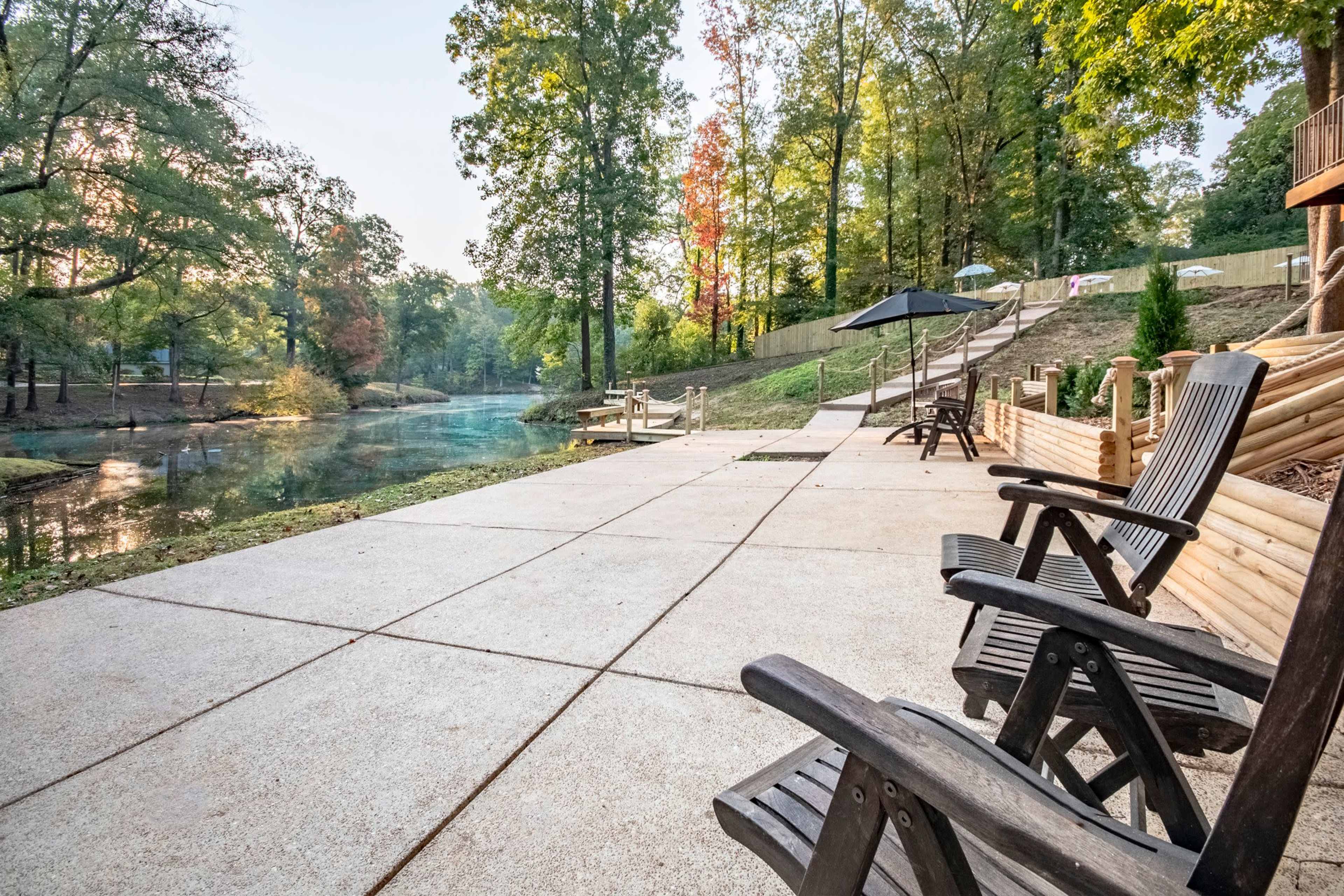 A tranquil riverside patio with wooden chairs and an umbrella, surrounded by trees and a clear waterway.