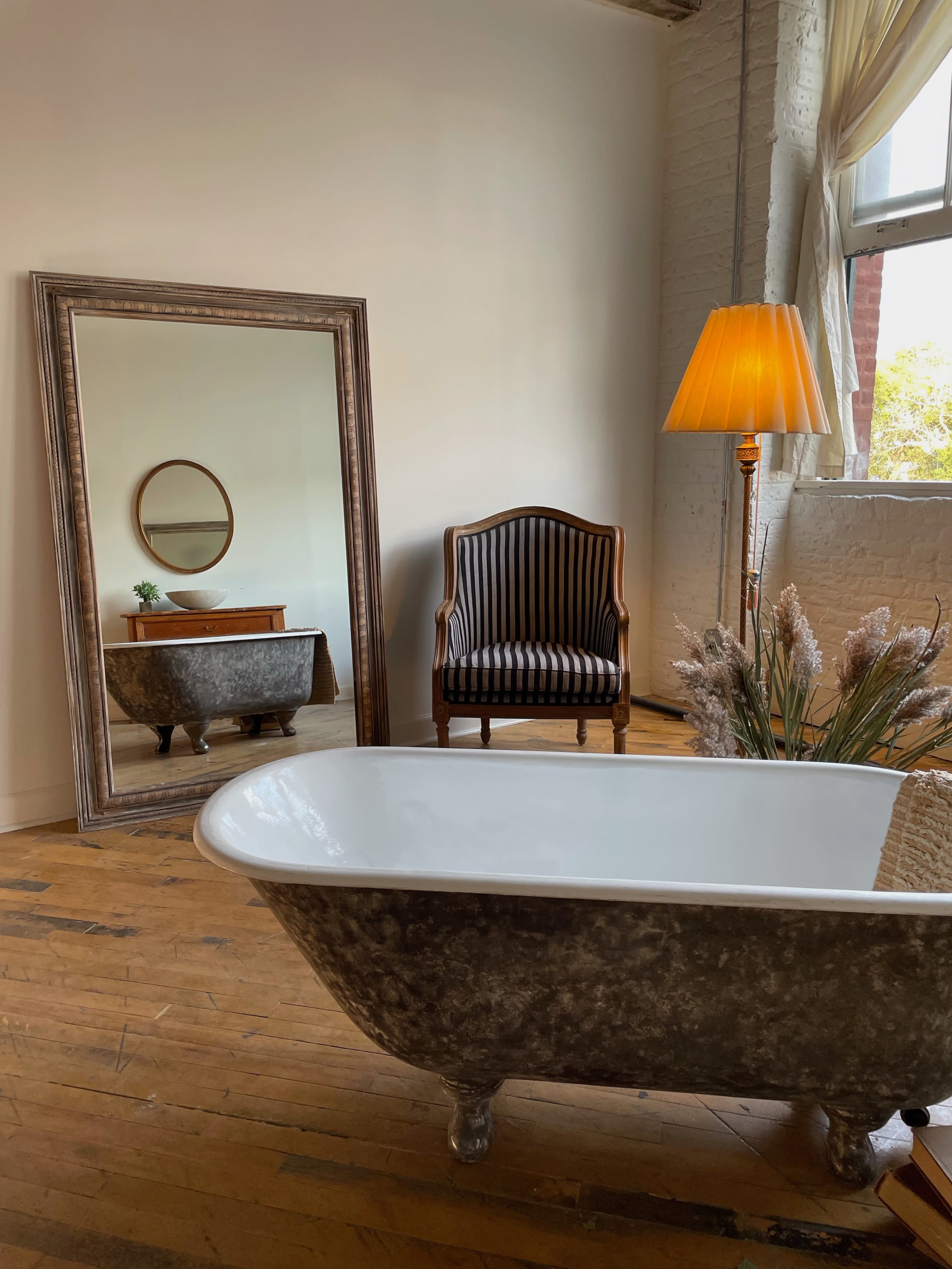 The scene features a vintage bathtub on wooden flooring, with a mirror reflecting a chair and a lamp beside a decorative plant.