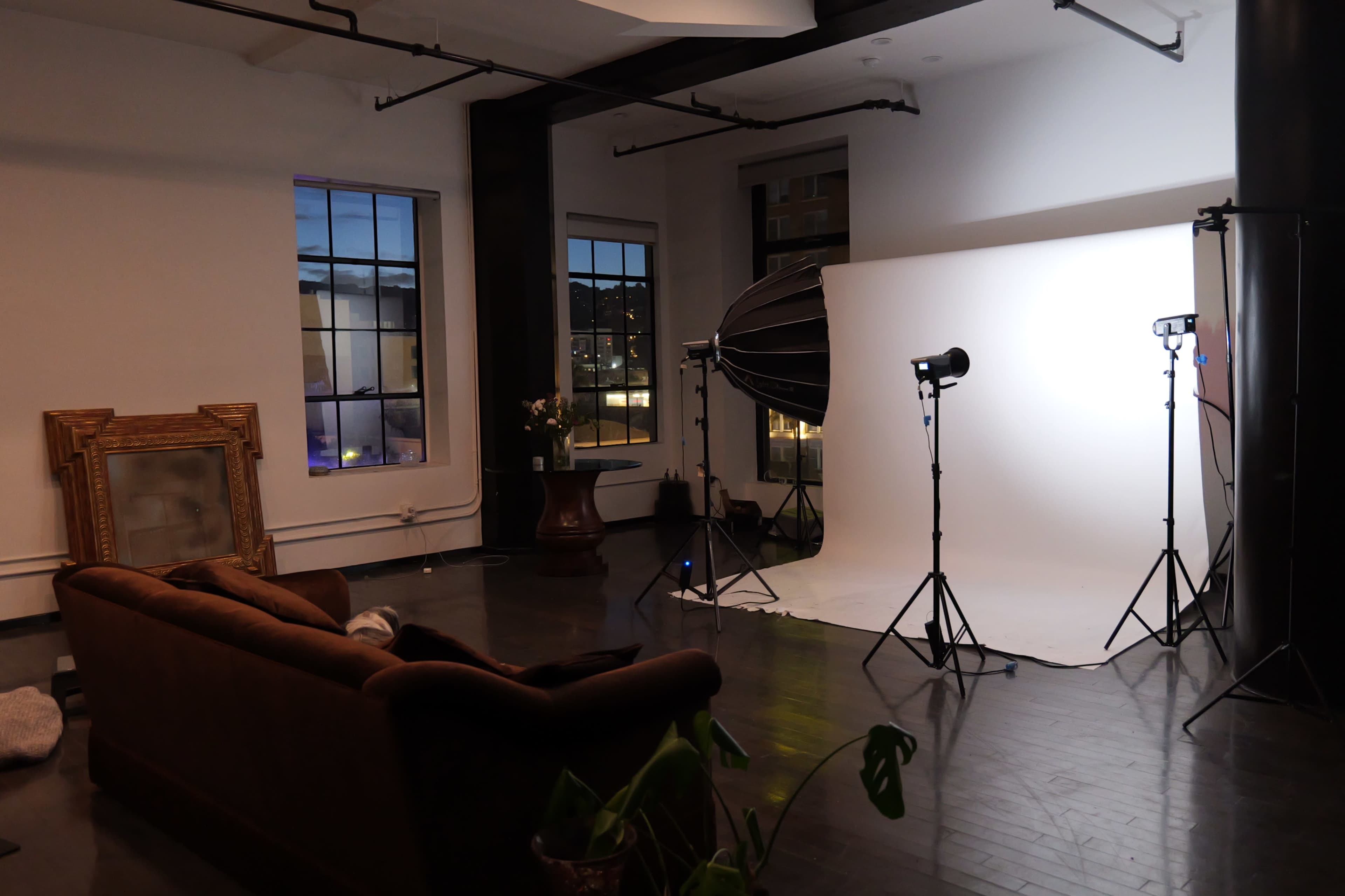 The scene features a spacious photo studio with a large white backdrop, lighting equipment, and a brown sofa against a wall with windows.