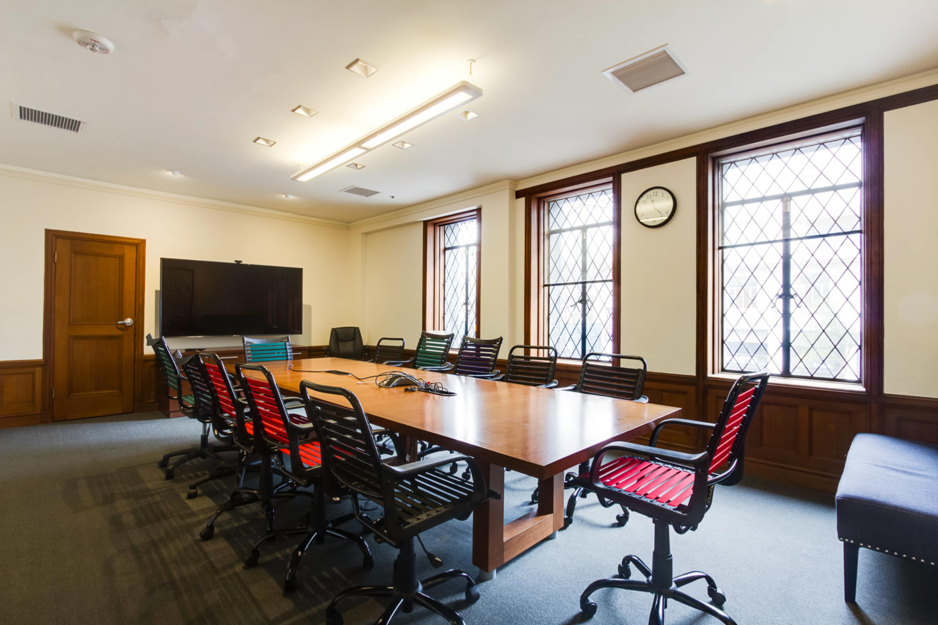 A conference room features a long wooden table surrounded by black chairs, with windows and a wall-mounted television.