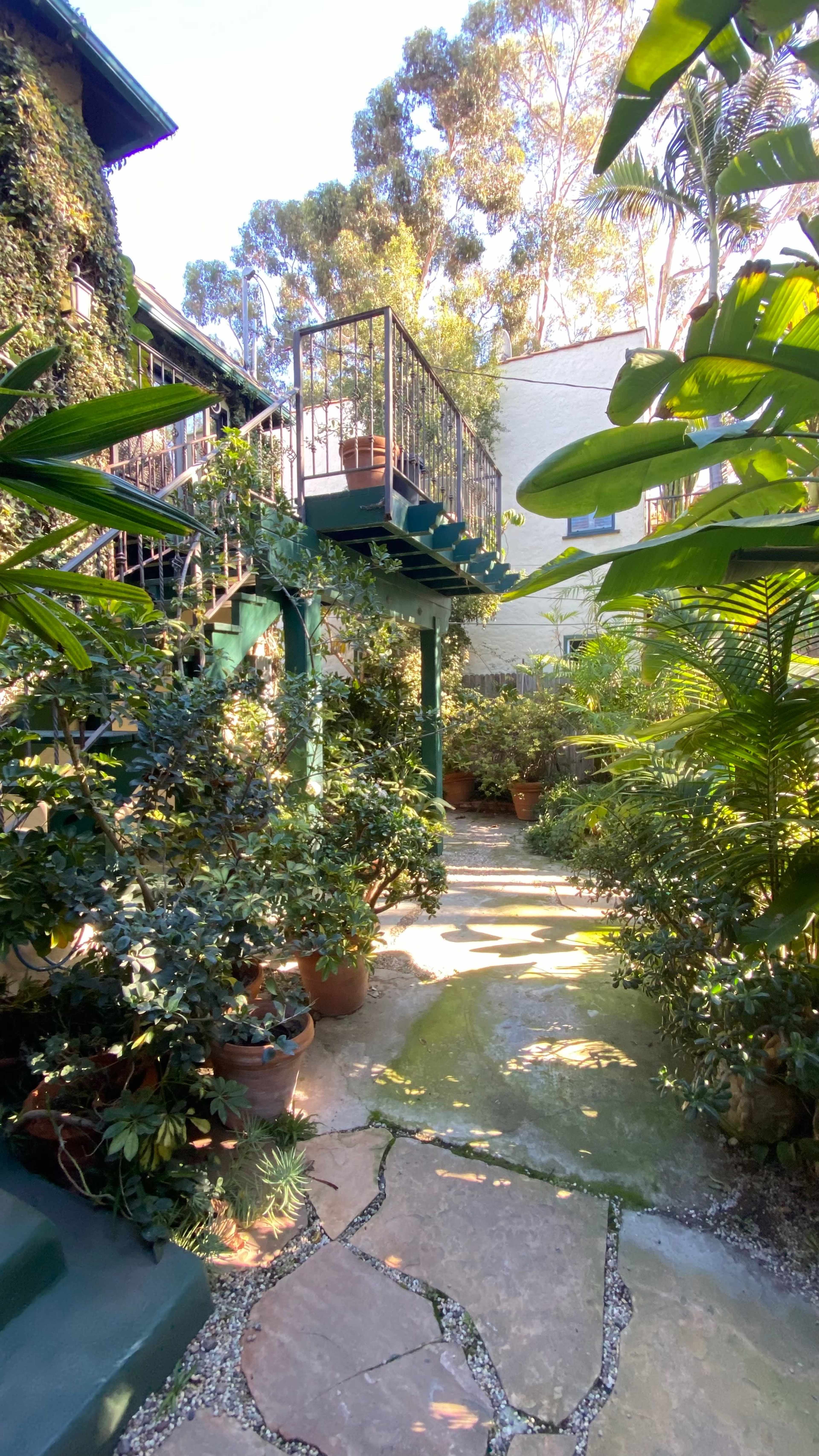 A stone pathway winding through a lush garden filled with potted plants and leading to a balcony above.