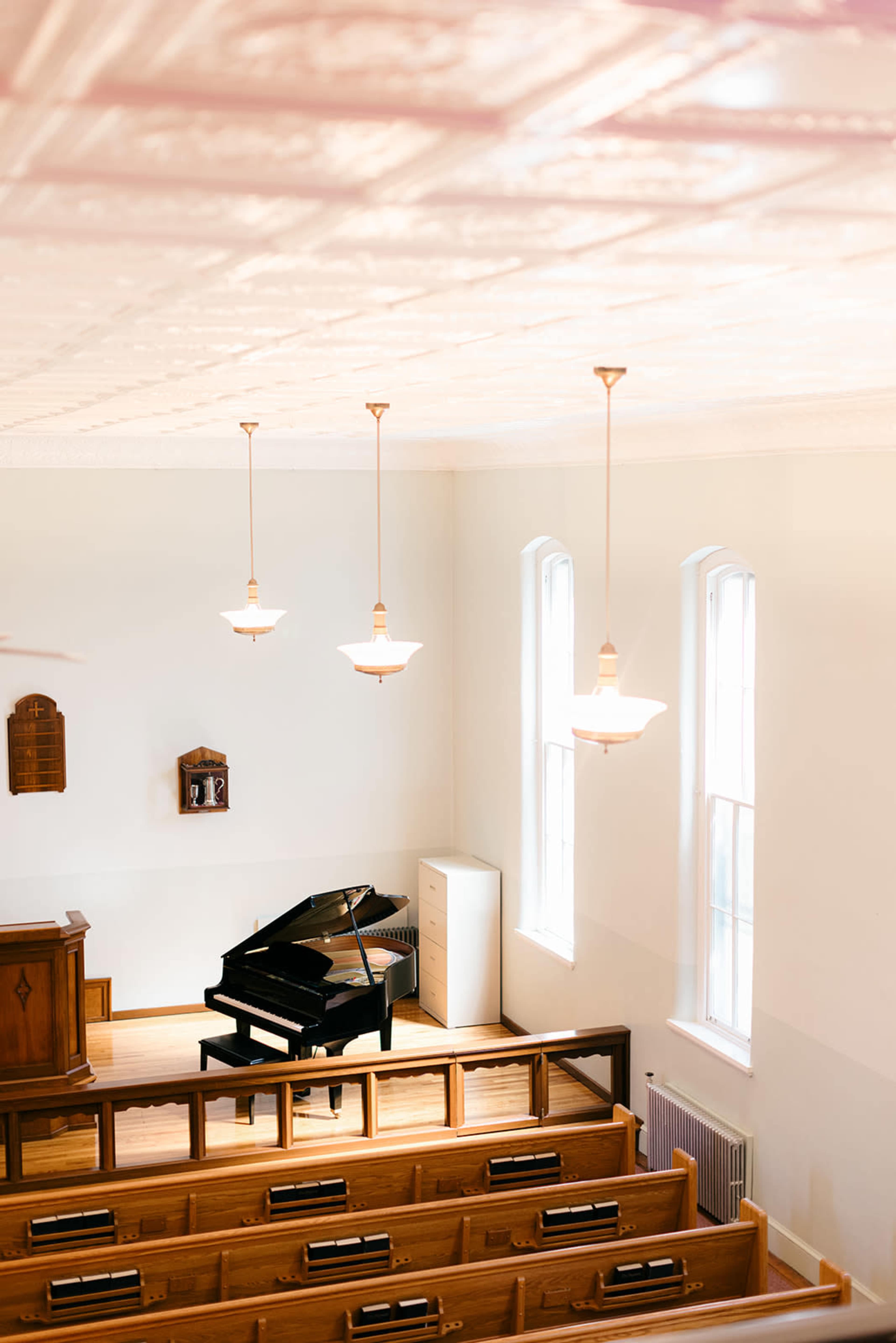 The image shows a view of an interior of a church with a grand piano prominently placed in front of a wooden podium, flanked by rows of wooden pews and large windows.