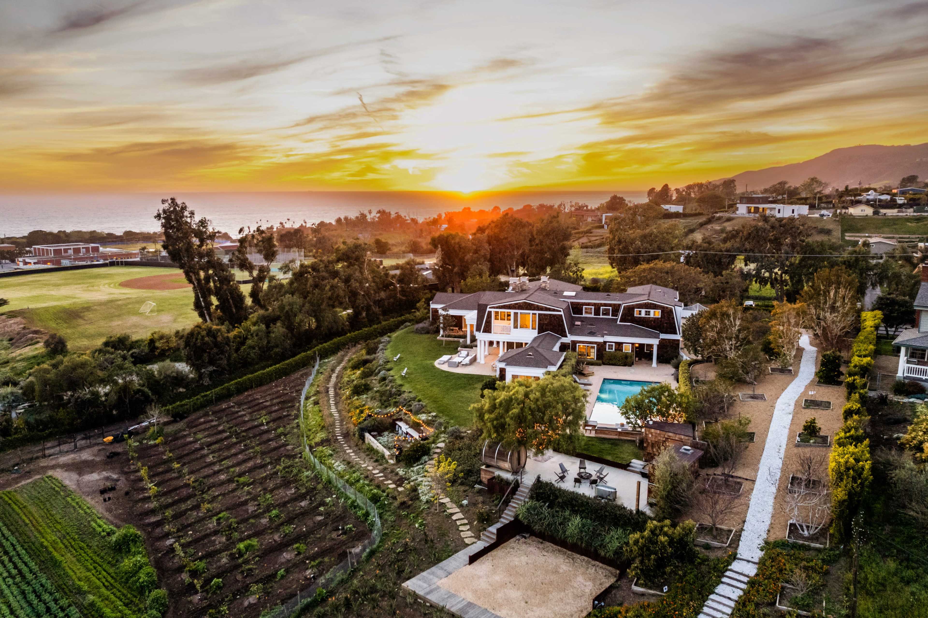 A modern house with a pool is surrounded by a well-maintained garden and farmland, overlooking a sunset by the ocean.