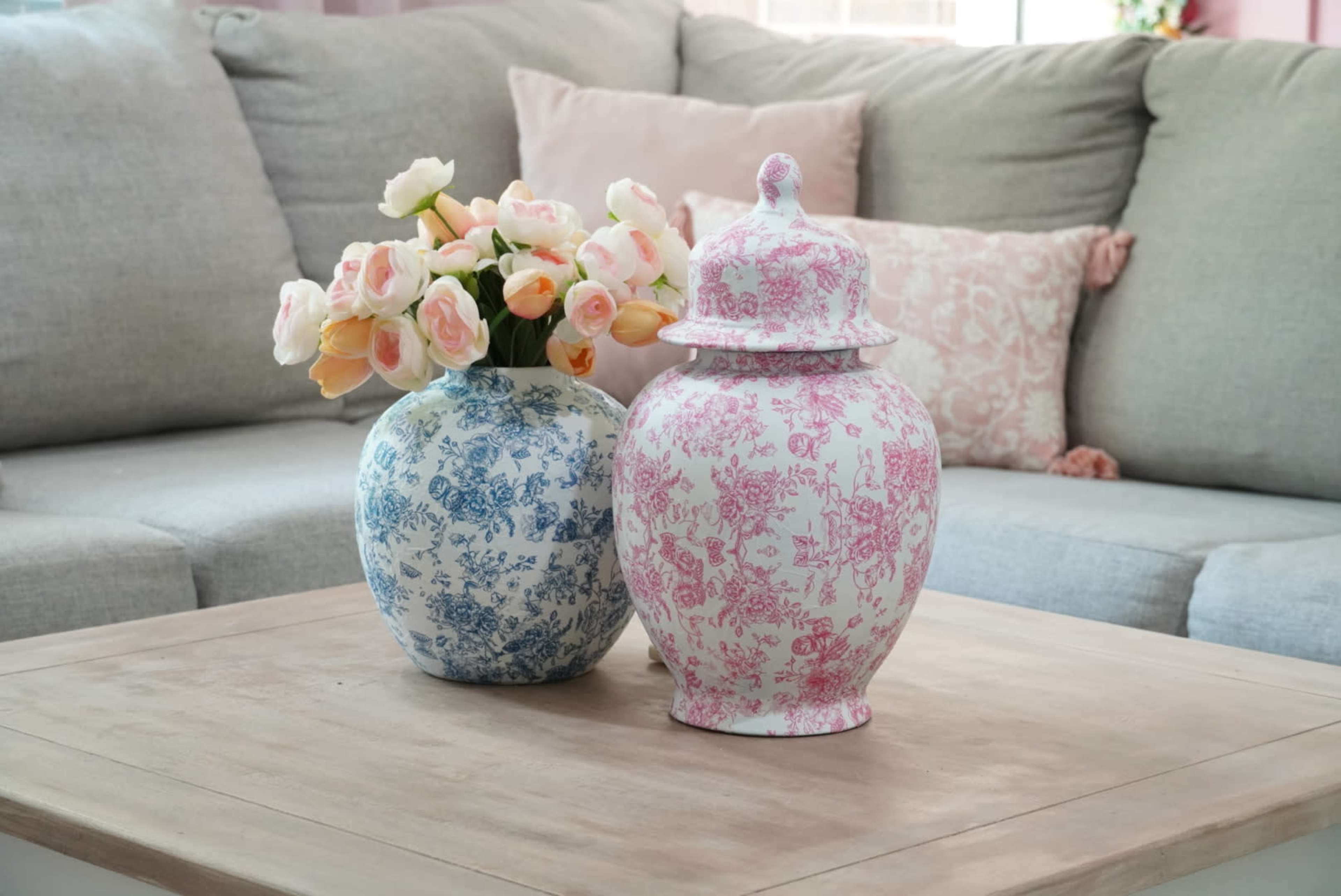 A blue floral vase and a pink floral lidded jar are placed on a wooden coffee table in front of a gray sofa.