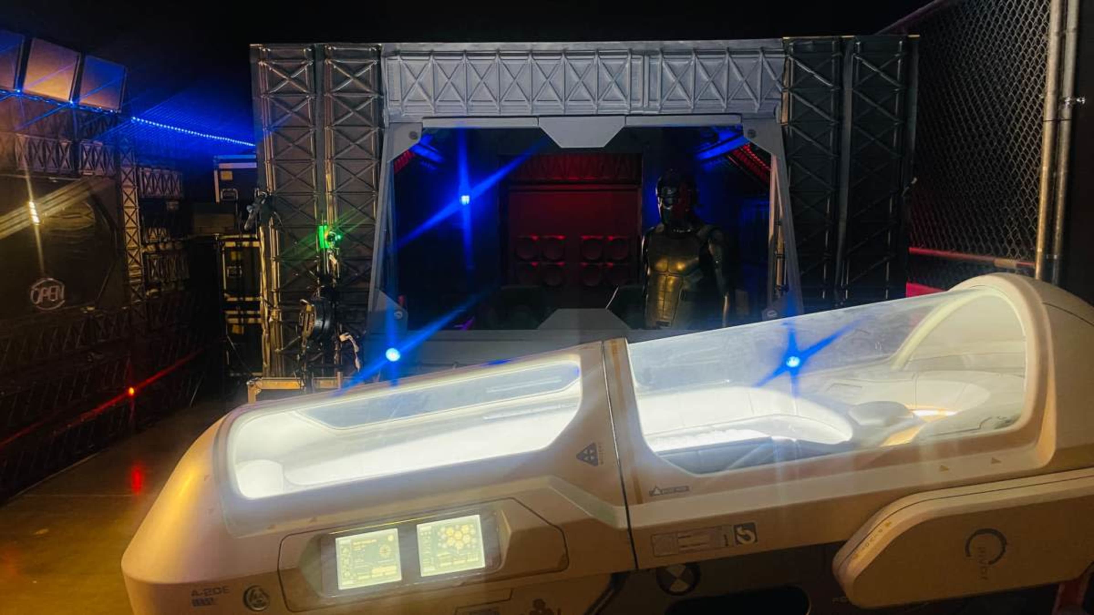 The image shows a futuristic set design featuring a sleek, white capsule with digital displays in the foreground, while a dark-colored figure stands in a shadowed background amid colorful lighting.