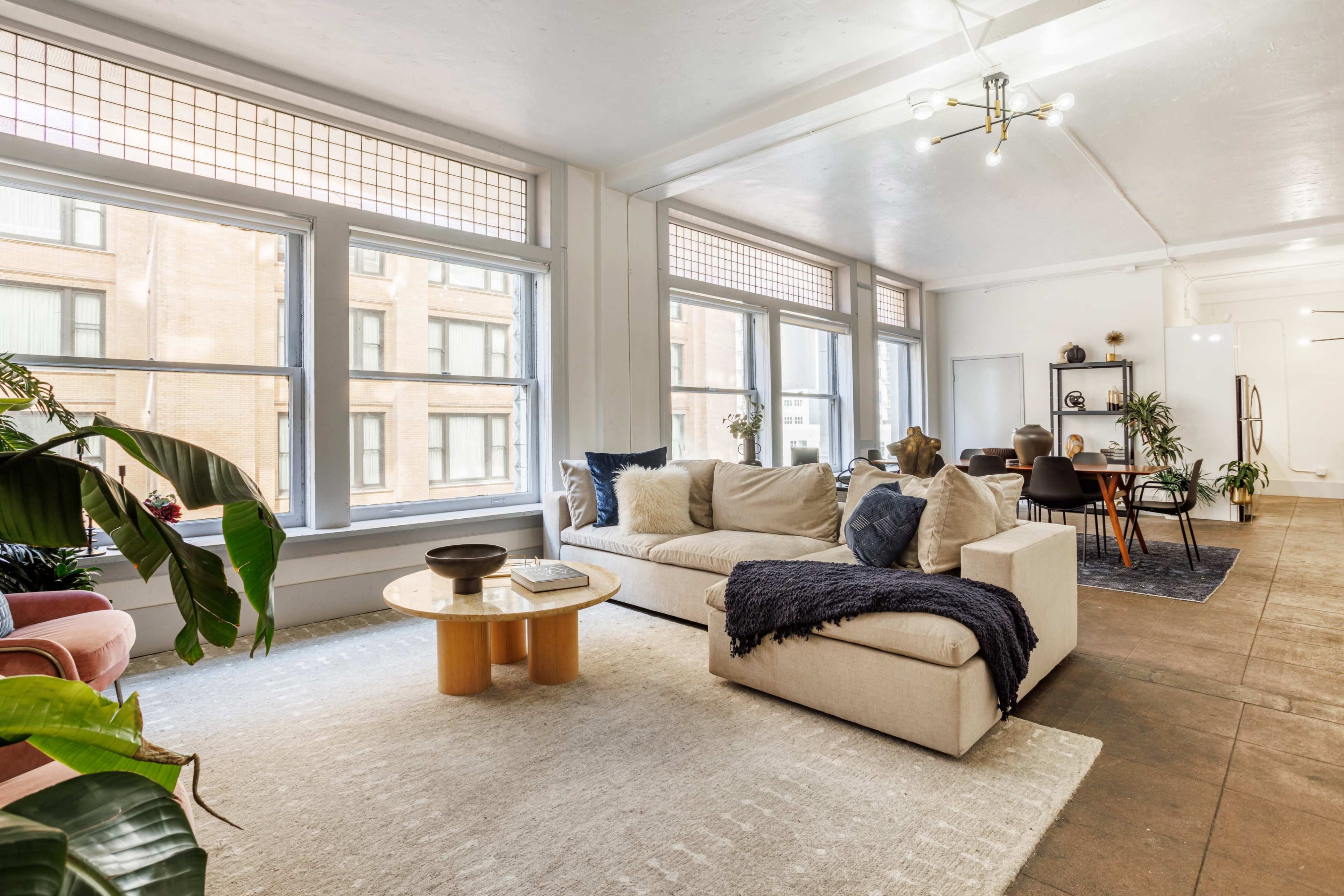 A modern living room features a cream sectional sofa, a round wooden coffee table, and large windows that offer natural light, with a dining area visible in the background.