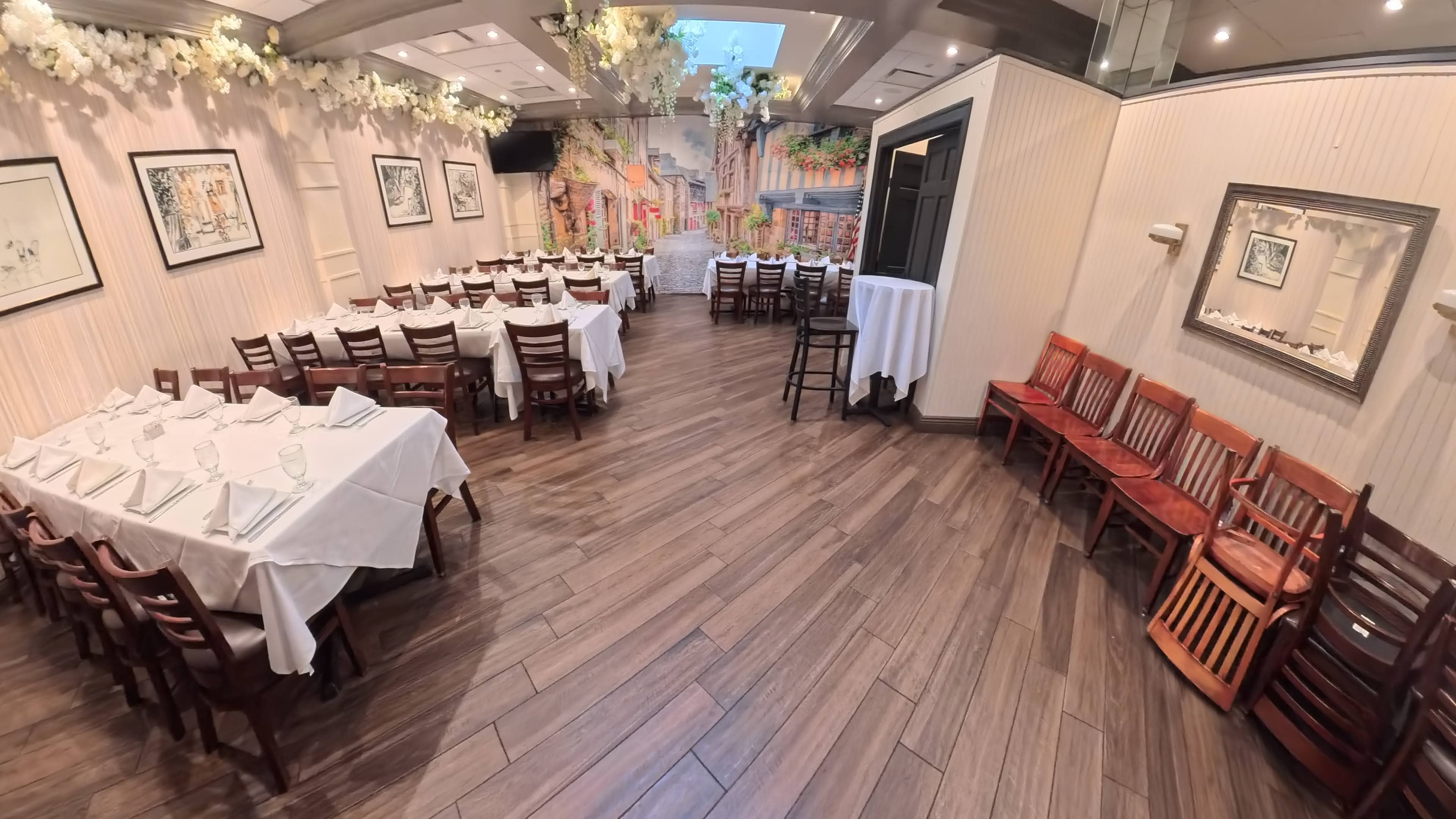 The image shows a restaurant interior featuring tables set with white tablecloths and wooden chairs arranged in a spacious dining area, along with a mural depicting a quaint street scene.