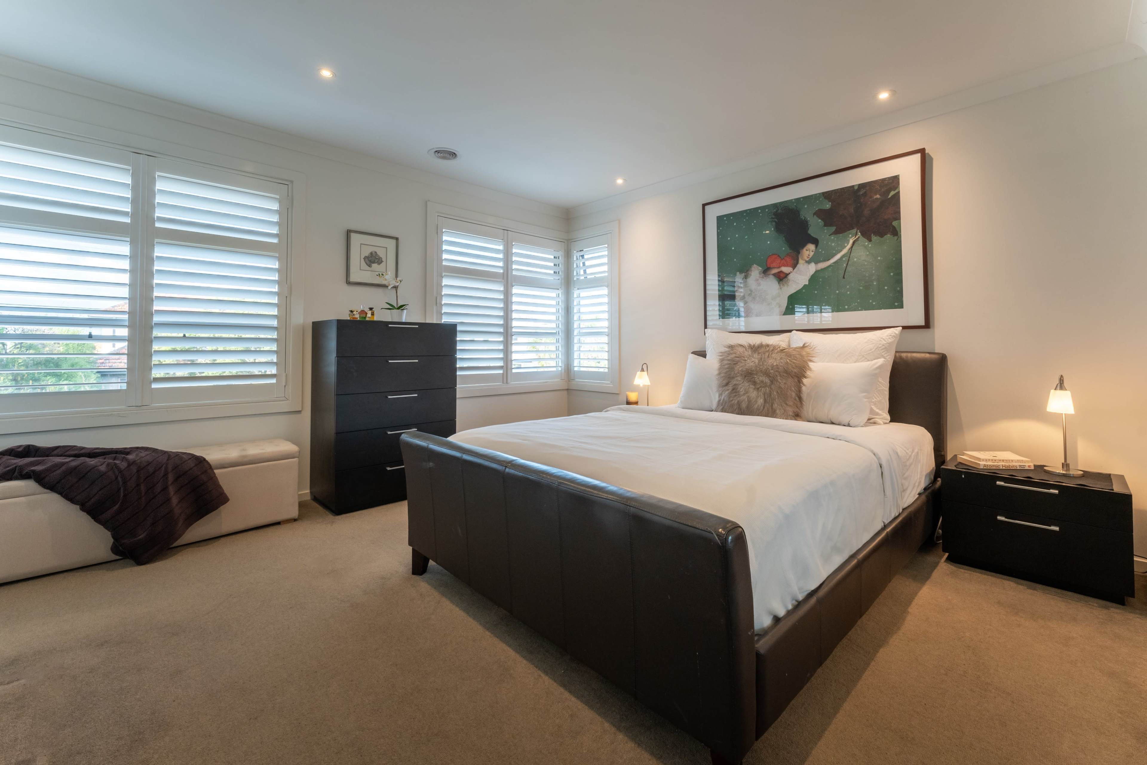 The image shows a well-lit bedroom featuring a bed with white linens, a black leather frame, a drawer cabinet, and large windows with slatted shutters.