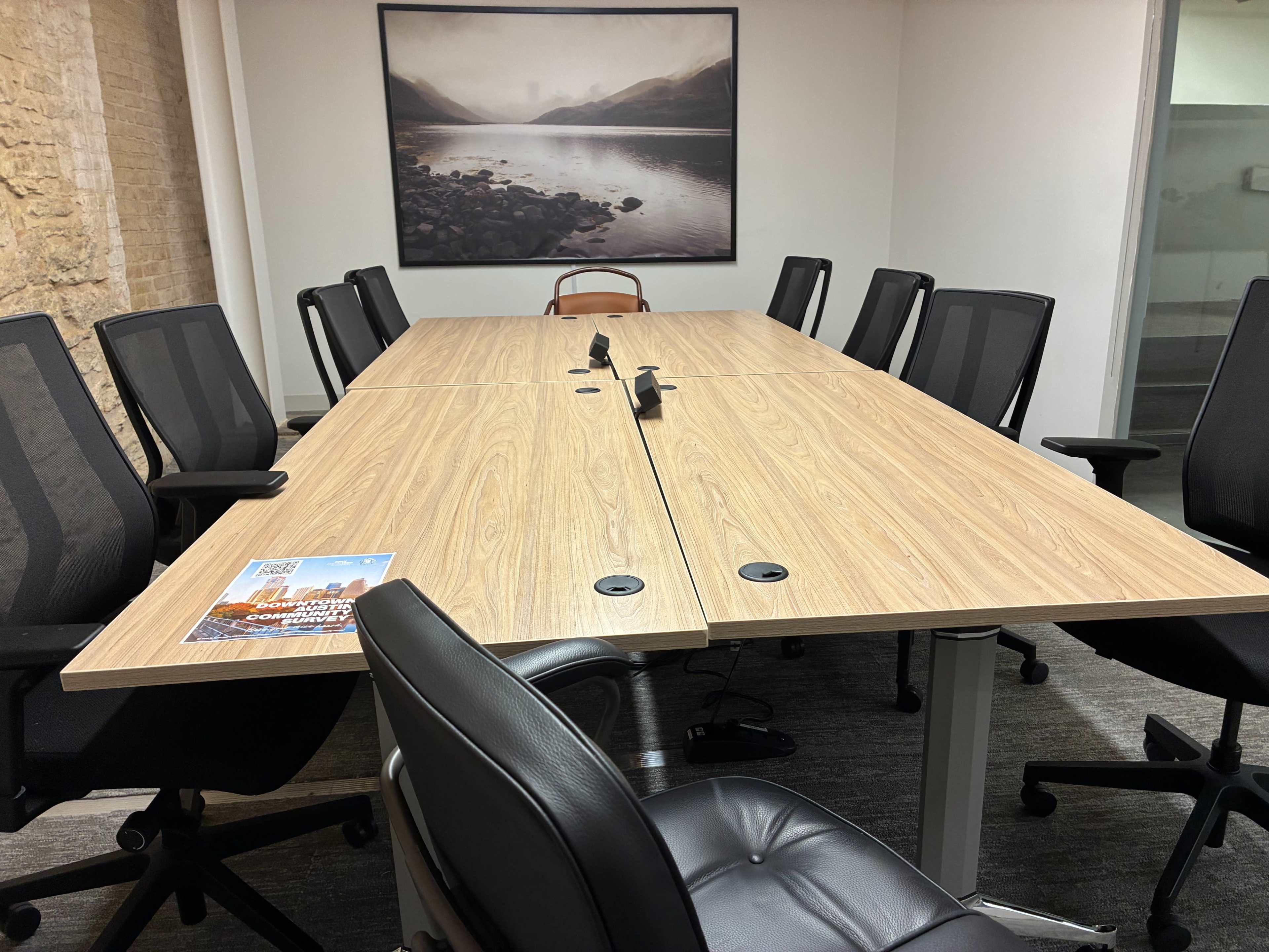 A long wooden conference table is set with ergonomic black chairs in a well-lit meeting room with a landscape photograph on the wall.