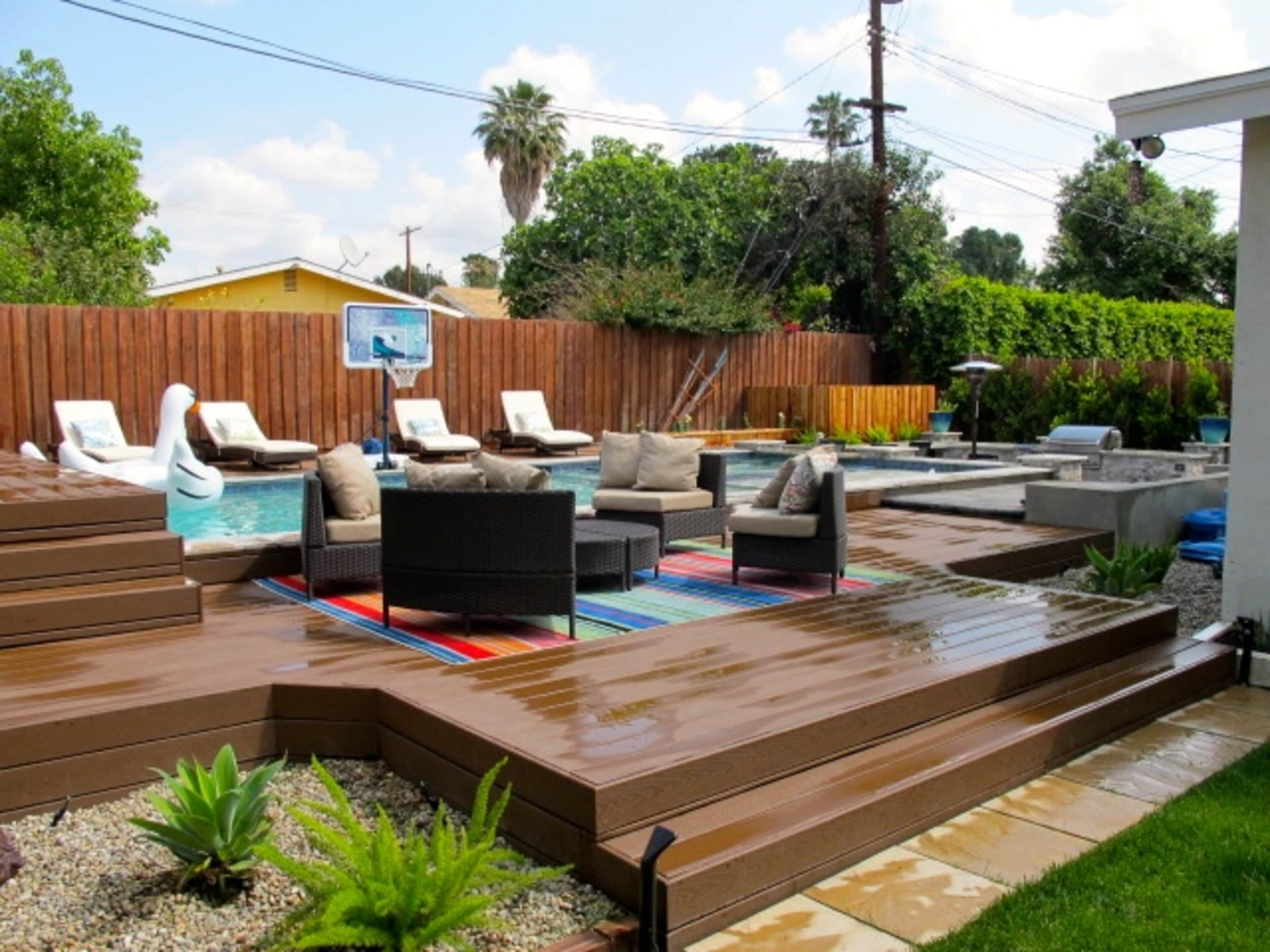 The image shows a residential backyard featuring a swimming pool surrounded by lounge chairs, a colorful rug, and landscaped areas with plants.