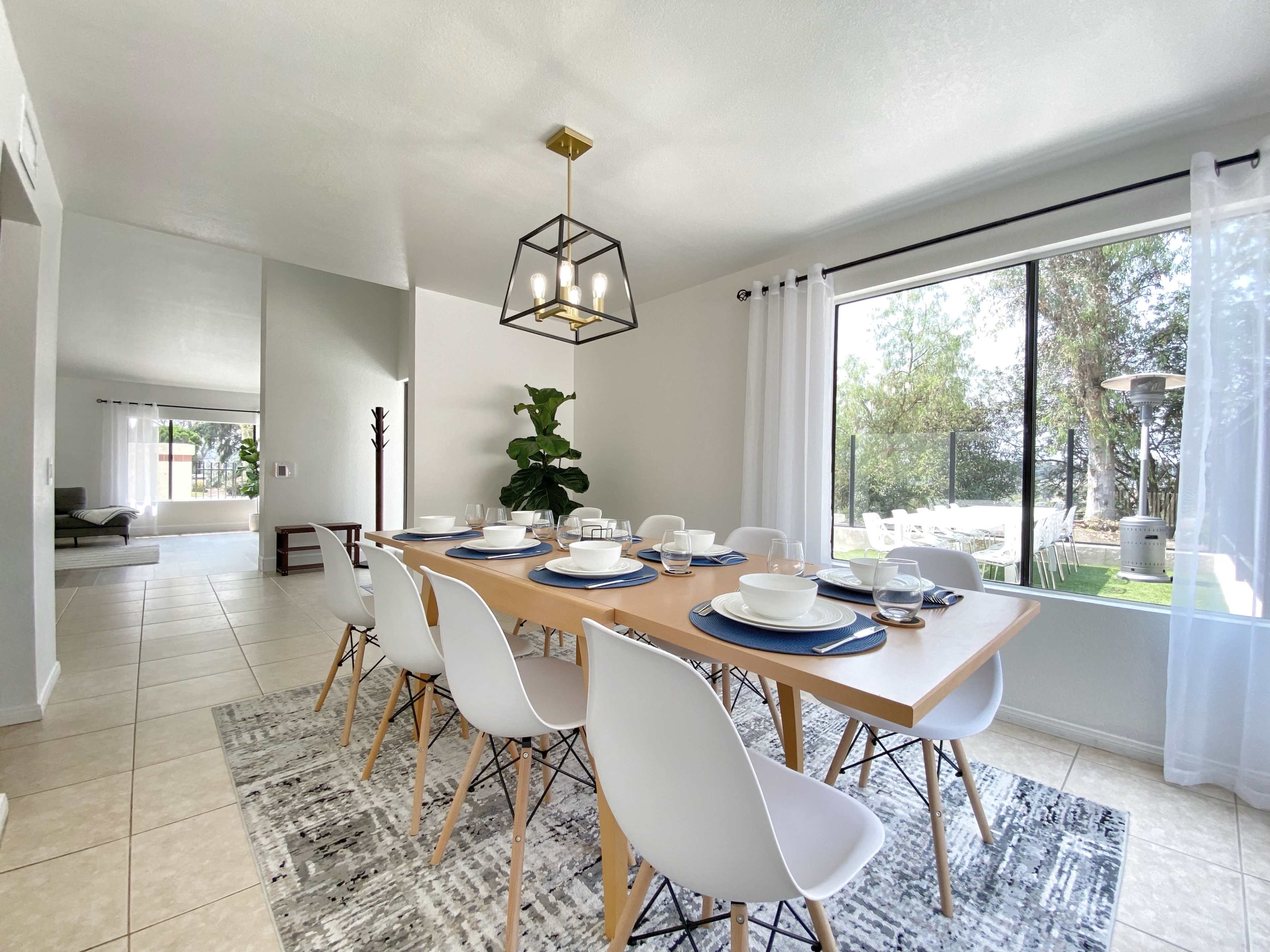A modern dining area features a long wooden table set for dinner, surrounded by white chairs, with large windows allowing natural light to illuminate the space.