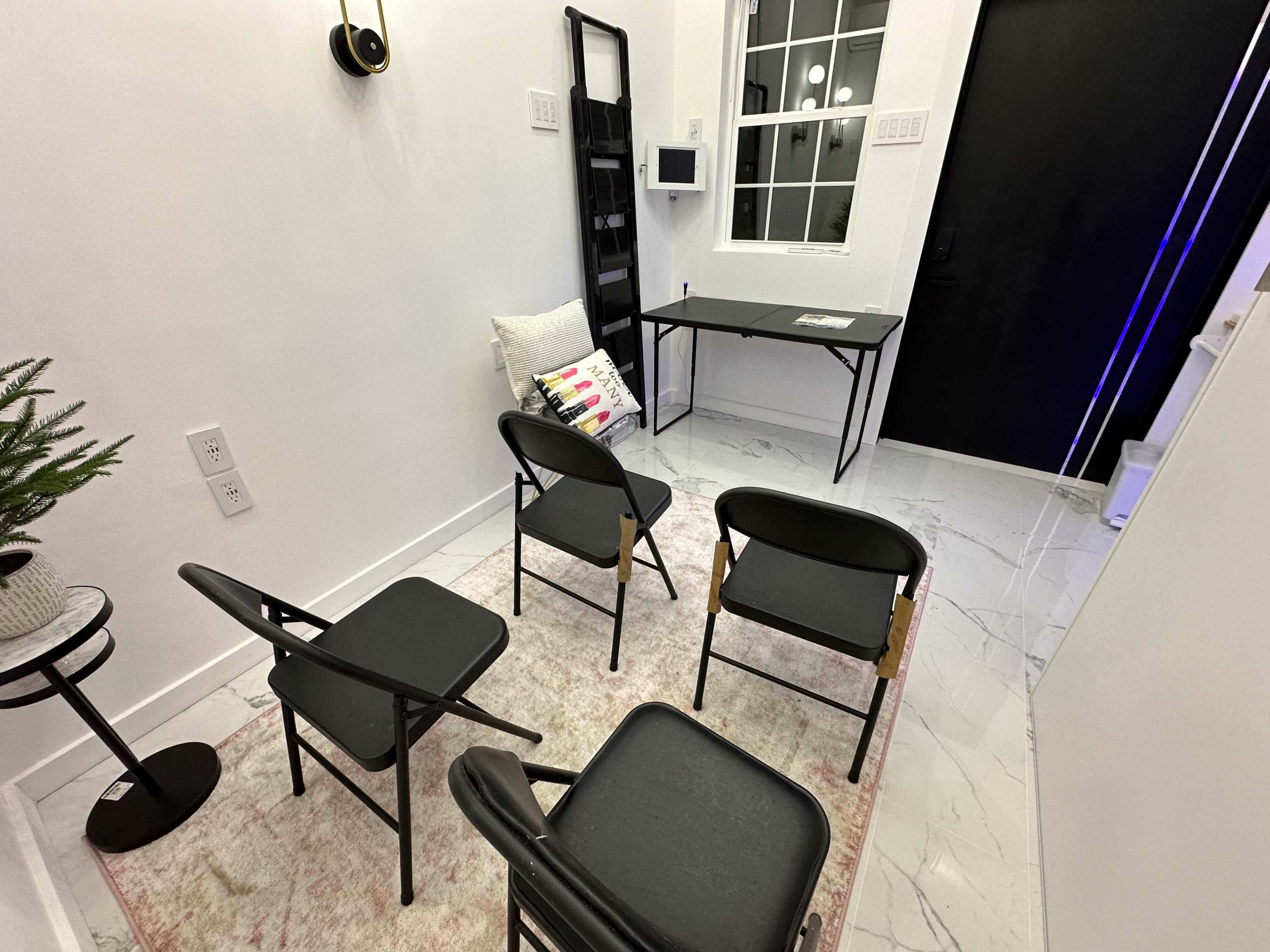 The image shows a small, minimalistic room with four black folding chairs arranged around a light-colored rug, alongside a table, a wall-mounted mailbox, and a plant in a pot.