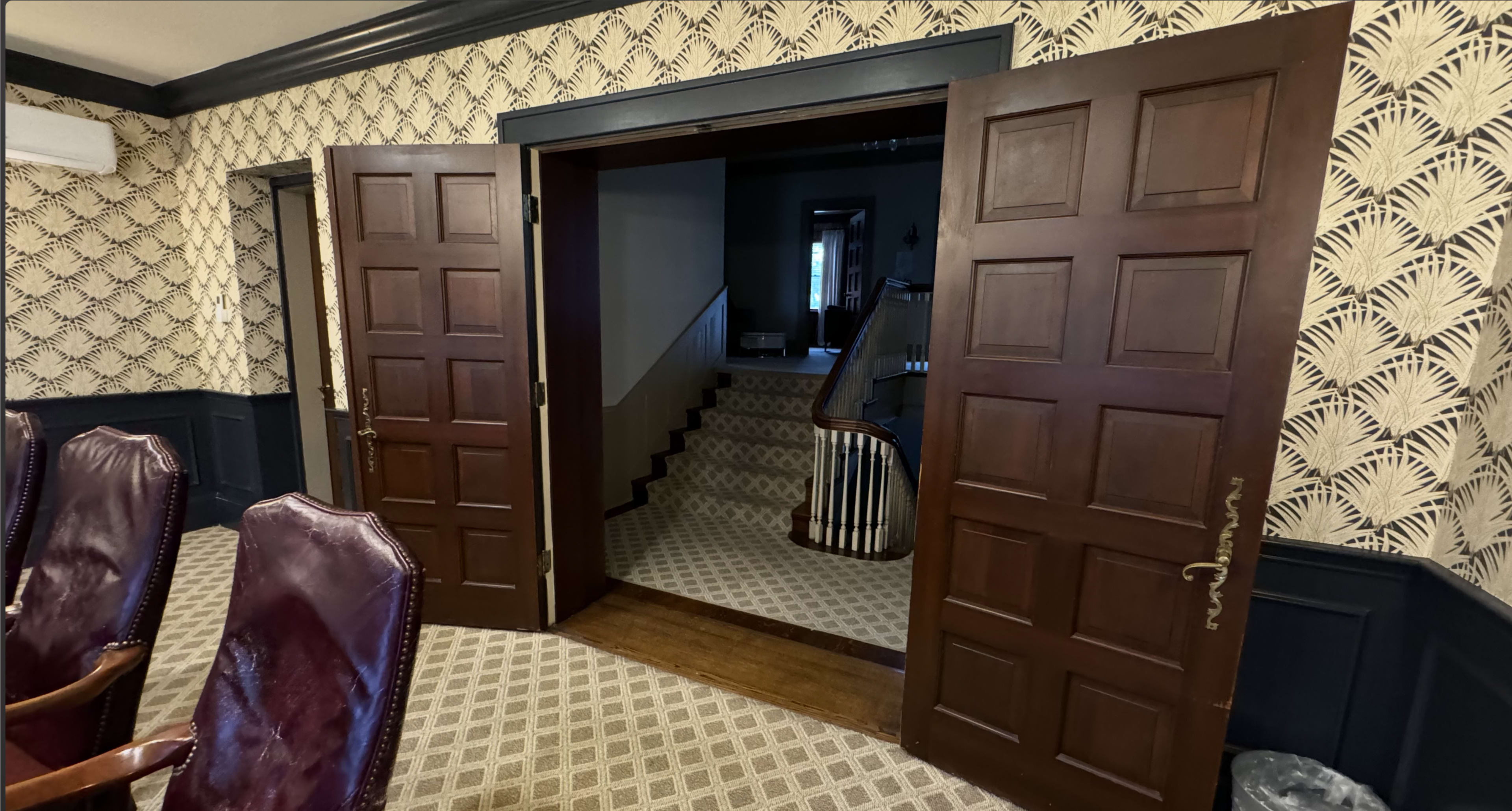 A pair of wooden doors opens to a staircase leading to a second floor, framed by patterned wallpaper and a carpeted floor.