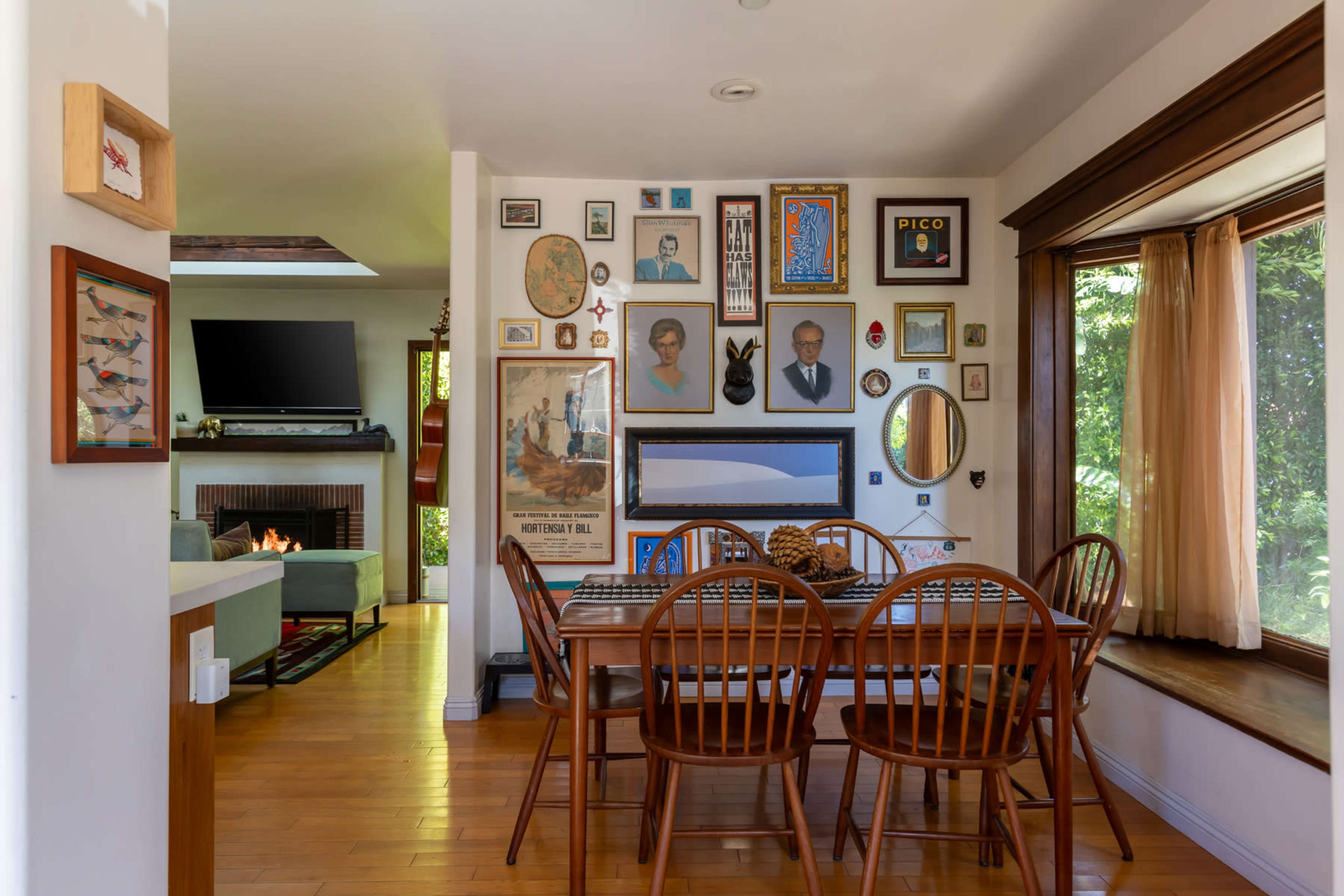 The image shows a dining area featuring a wooden table surrounded by chairs, with a gallery wall adorned with various framed artworks and photographs.