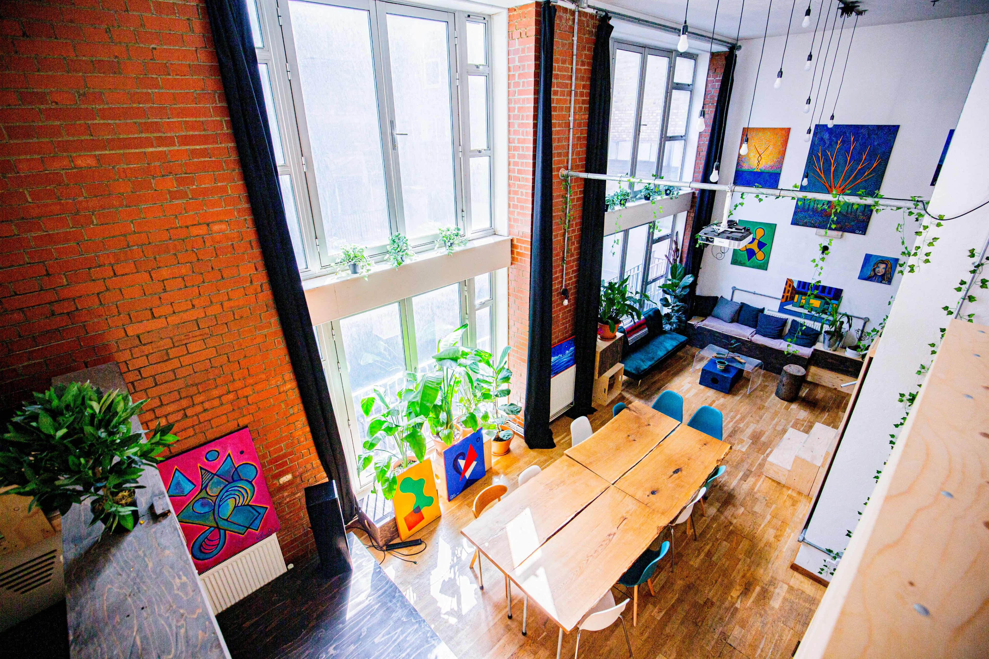 The image shows a spacious room with large windows, a long wooden table, colorful artwork on the walls, and a seating area filled with plants.