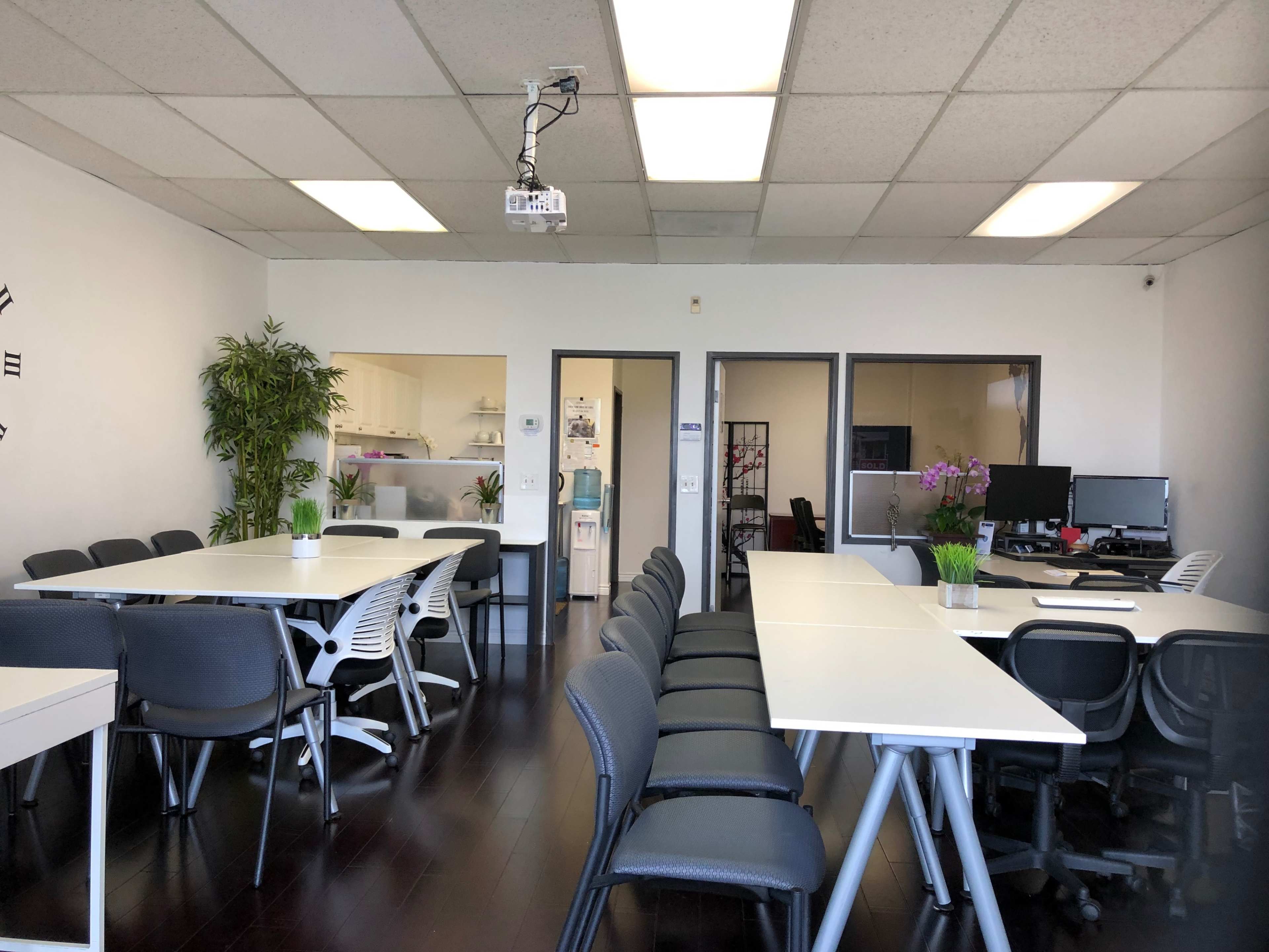 A modern office space with several white tables and chairs, a projector hanging from the ceiling, and a kitchenette visible through glass doors.