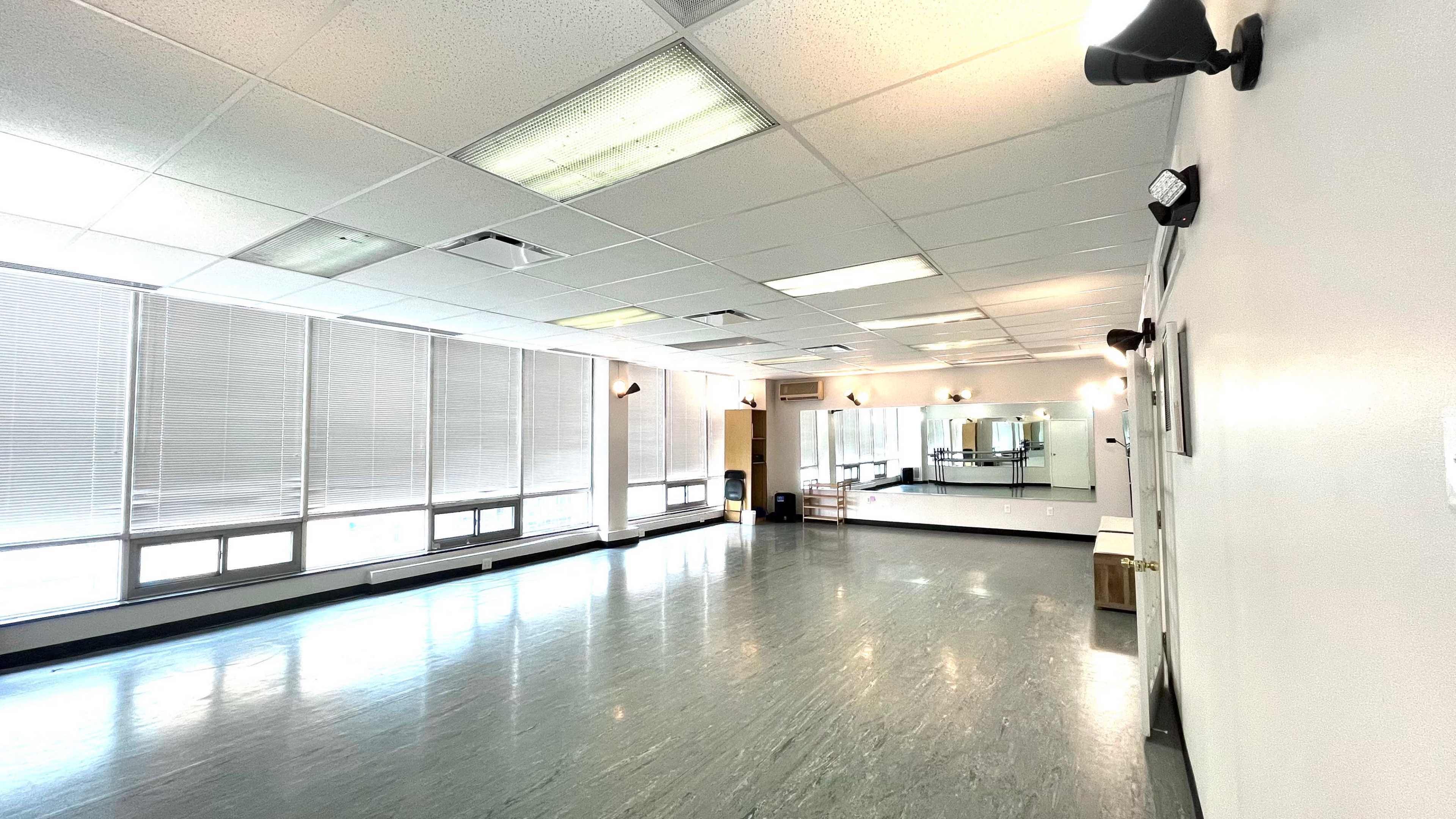 The image shows a spacious, well-lit studio with large windows and a mirrored wall, featuring a smooth floor and minimal furnishings.