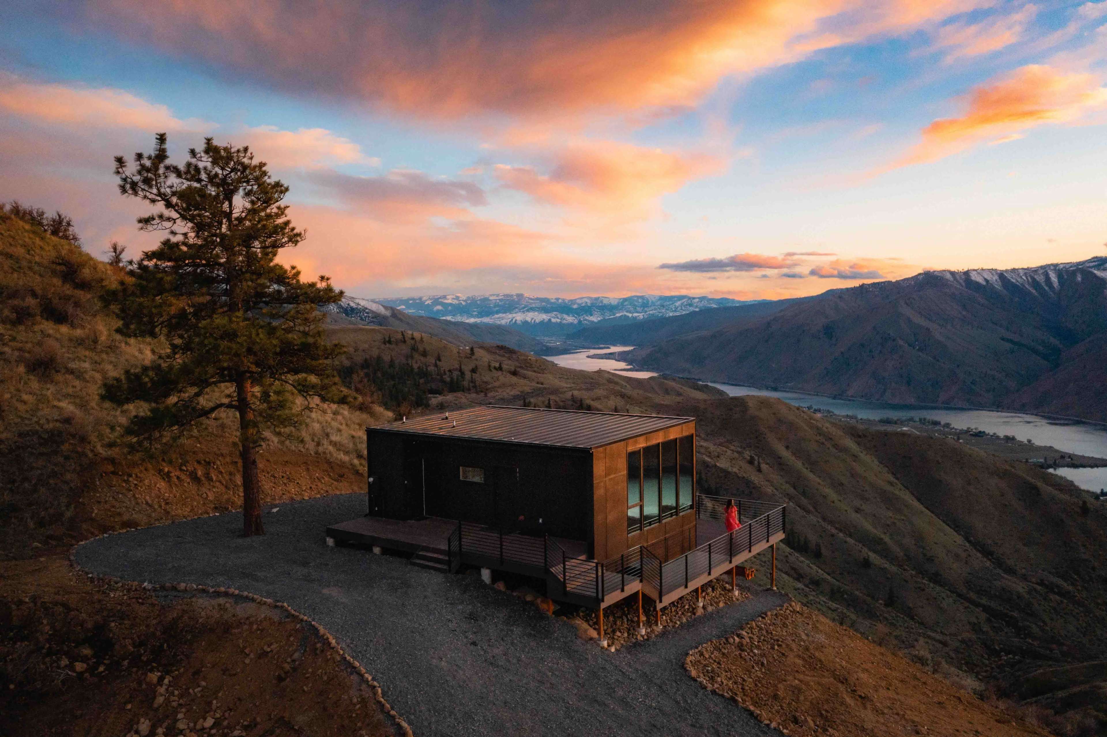 A modern cabin sits on a hillside overlooking a lake, with mountains in the background and a person standing near the edge.
