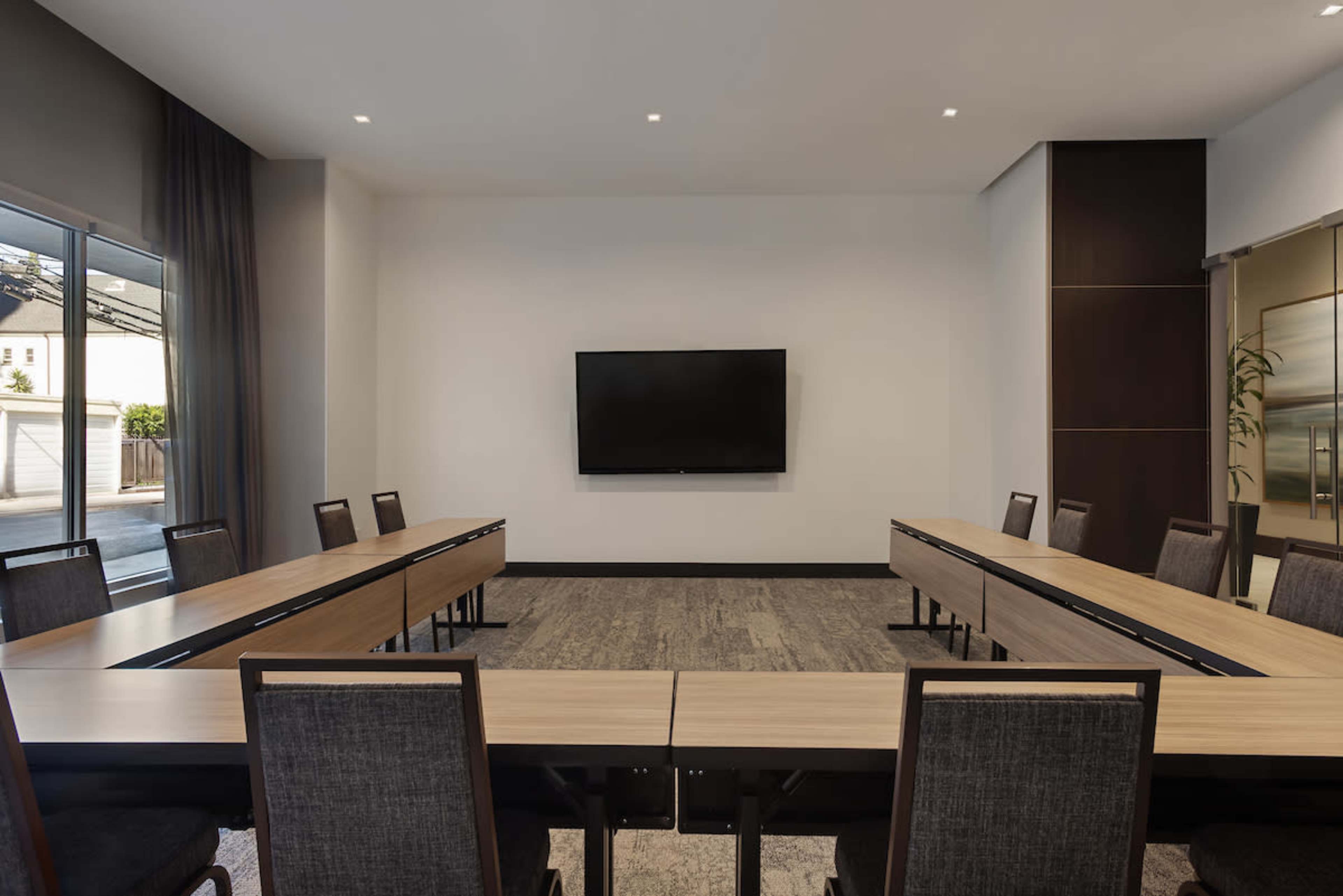The image shows a modern conference room arranged with multiple tables and chairs, facing a large wall-mounted television.