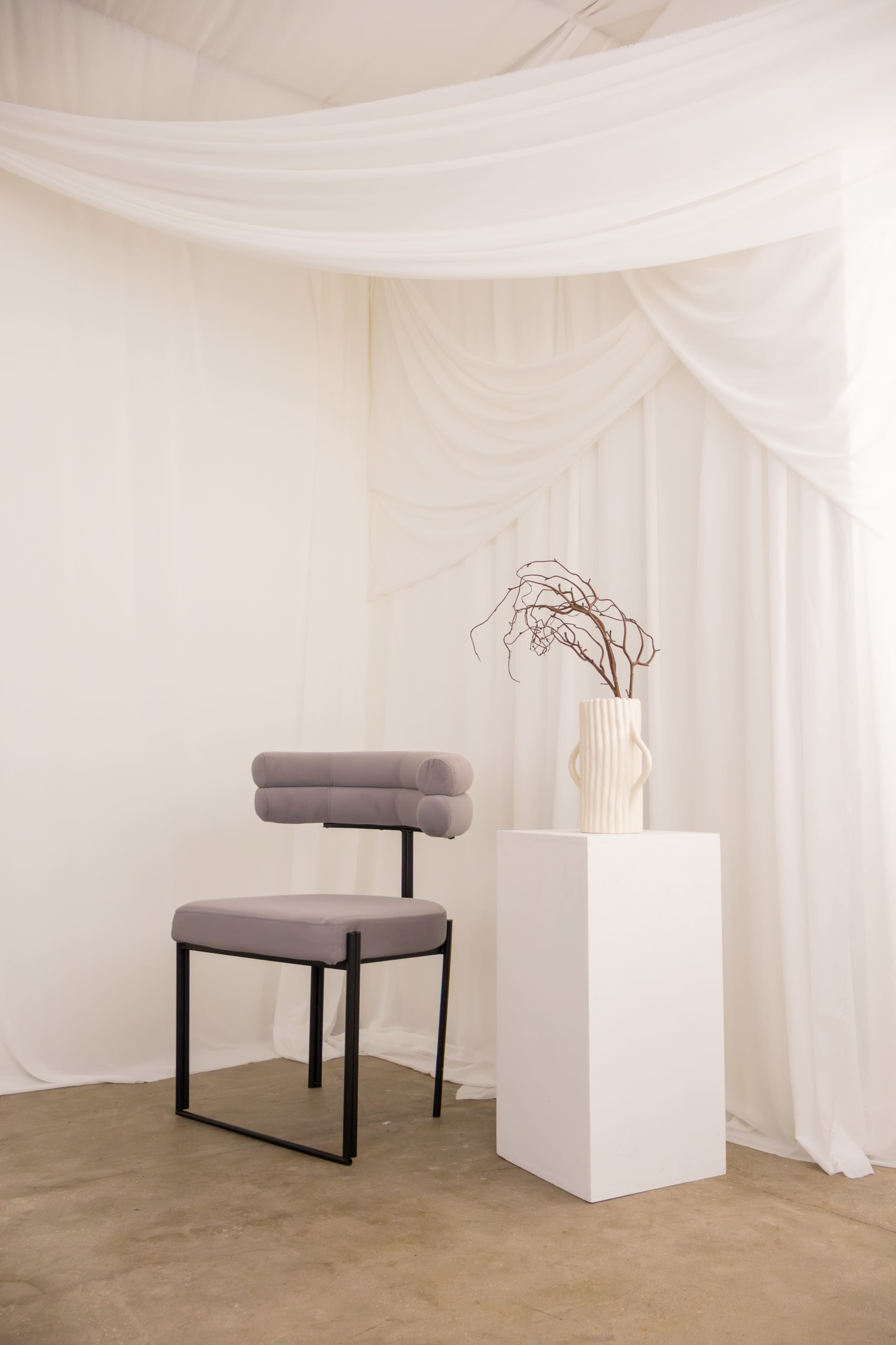 A modern gray chair is positioned next to a white pedestal displaying a vase with branches, all set against a backdrop of flowing white fabric.
