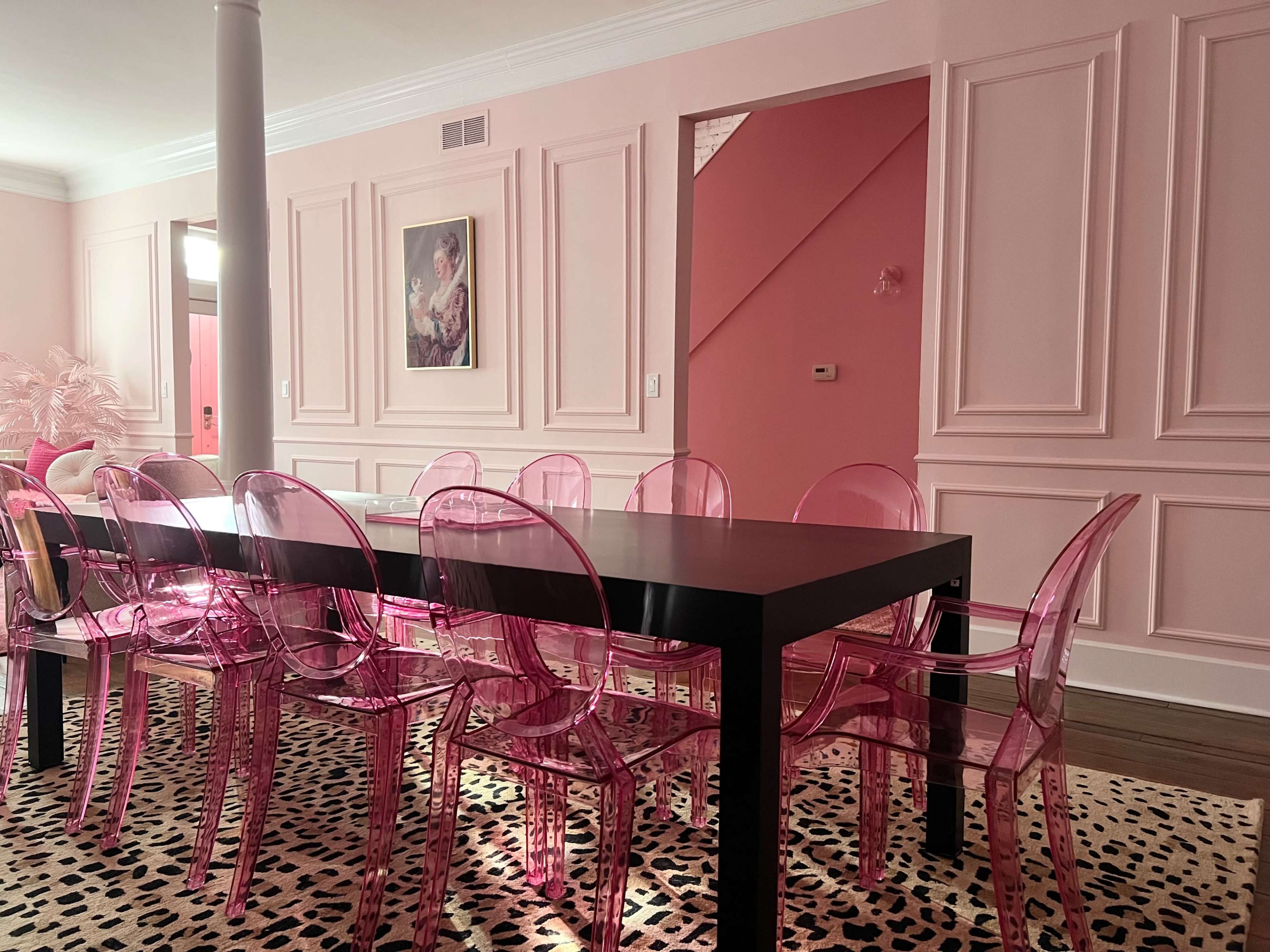A rectangular black dining table surrounded by transparent pink chairs sits on a leopard-print rug in a room with pink walls and wainscoting.