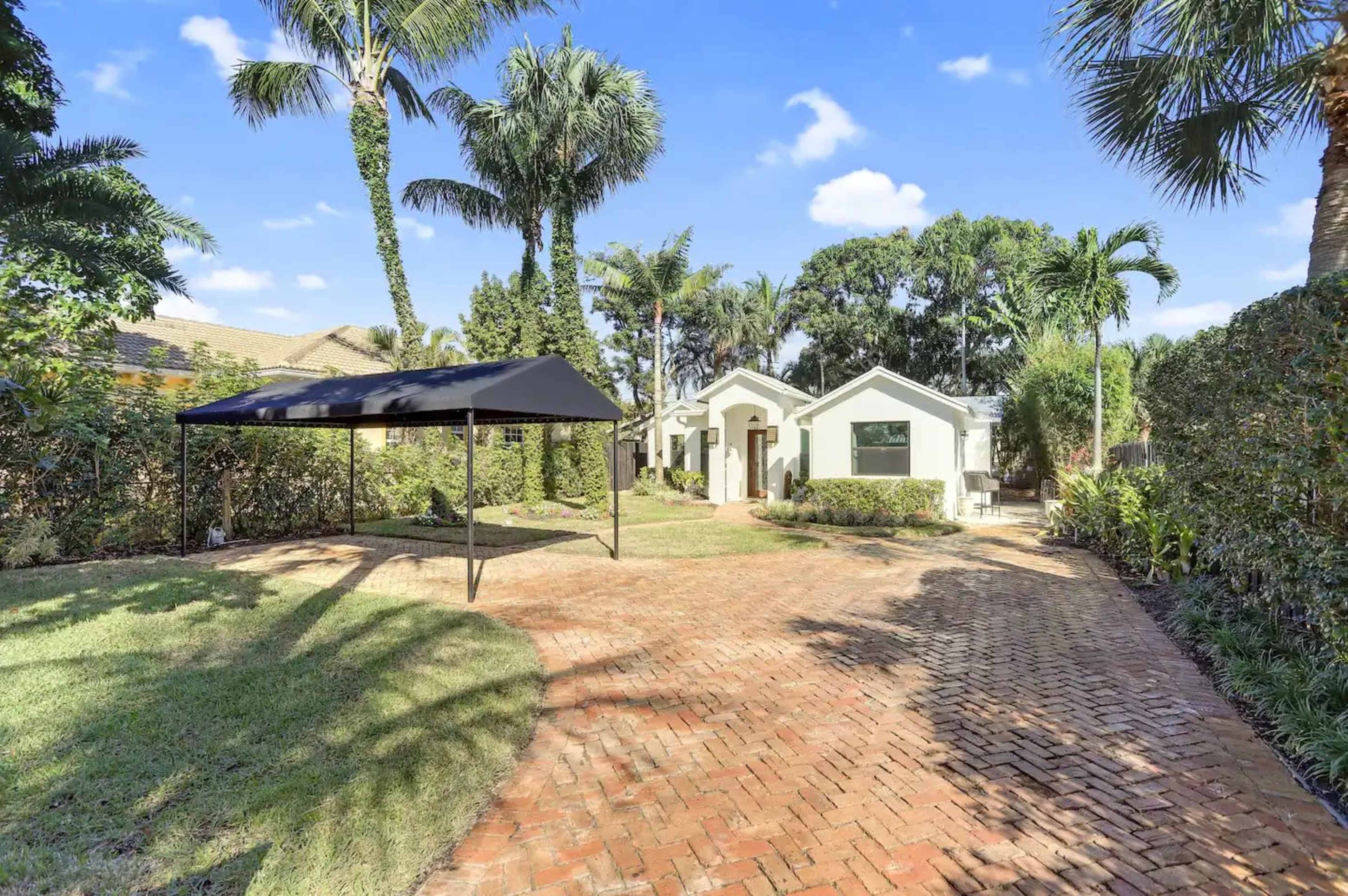 The image shows a landscaped backyard with a brick patio, palm trees, and a small canopy near a white house.
