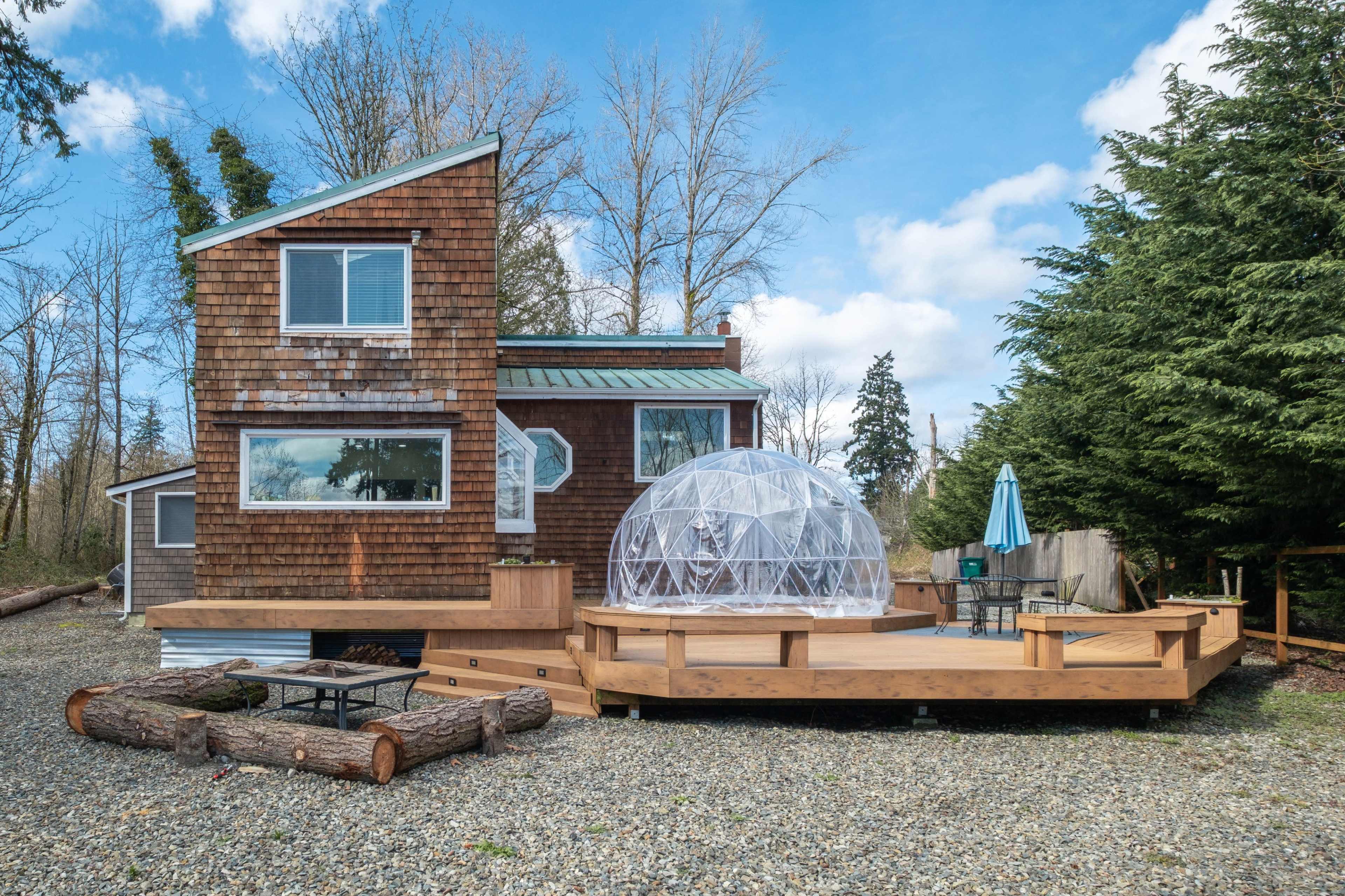 A wooden house with a green roof features a large deck and a transparent dome structure on the patio, surrounded by trees and gravel.