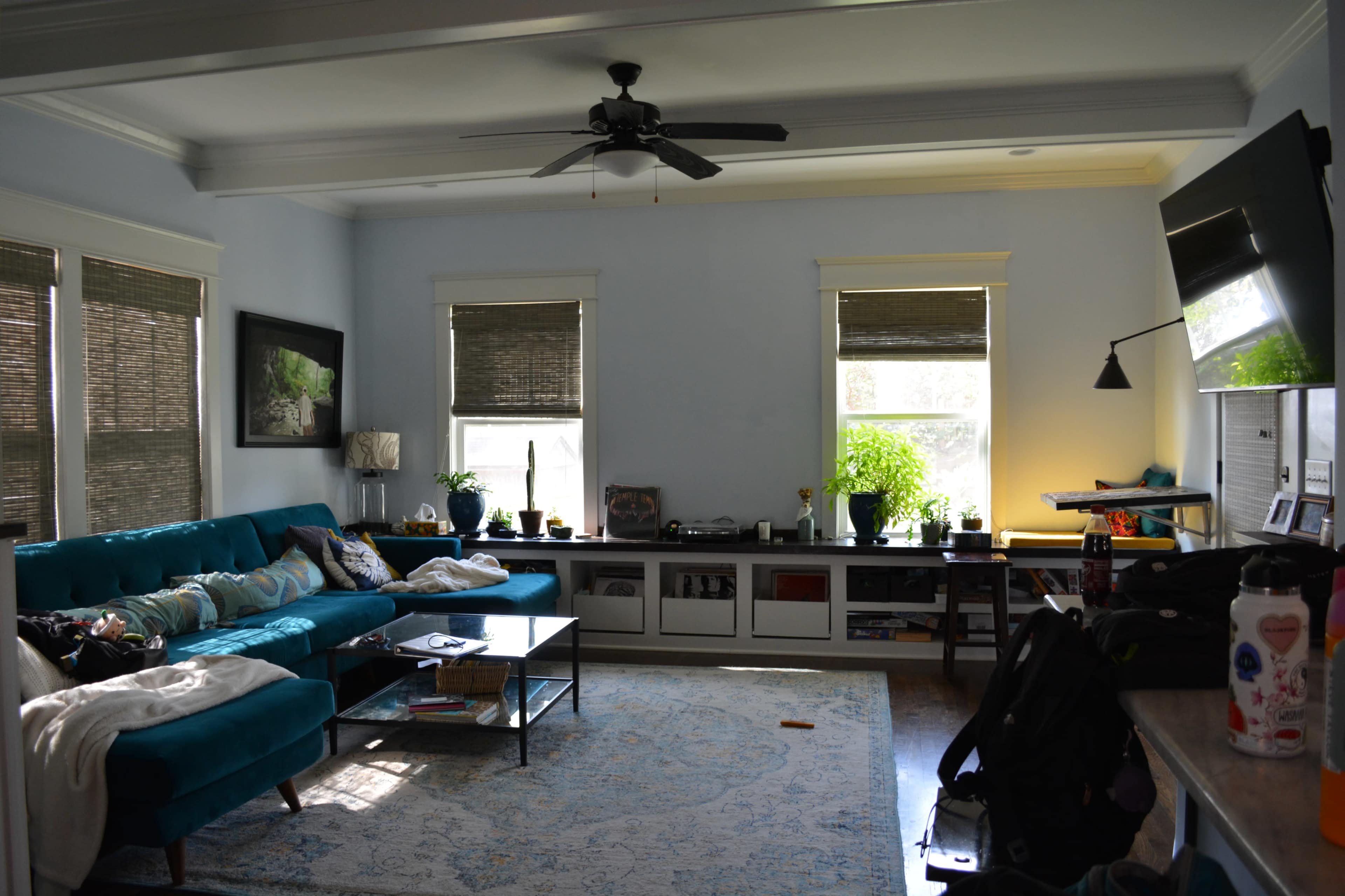 A spacious living room features a teal sofa, a coffee table, large windows with shades, and a TV mounted on the wall.