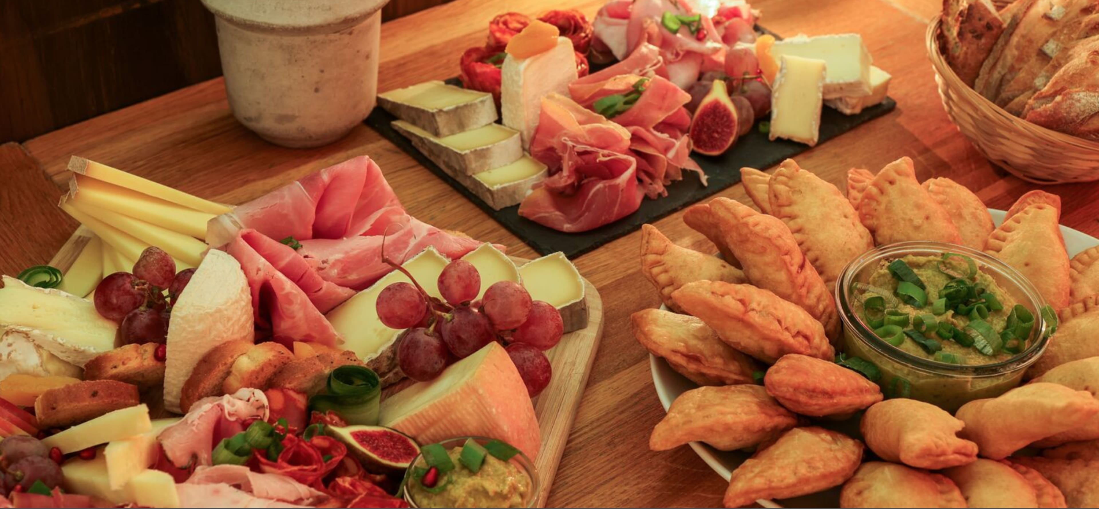 A wooden table displays an assortment of cheeses, cured meats, fresh fruit, and fried pastries arranged on platters and a basket.