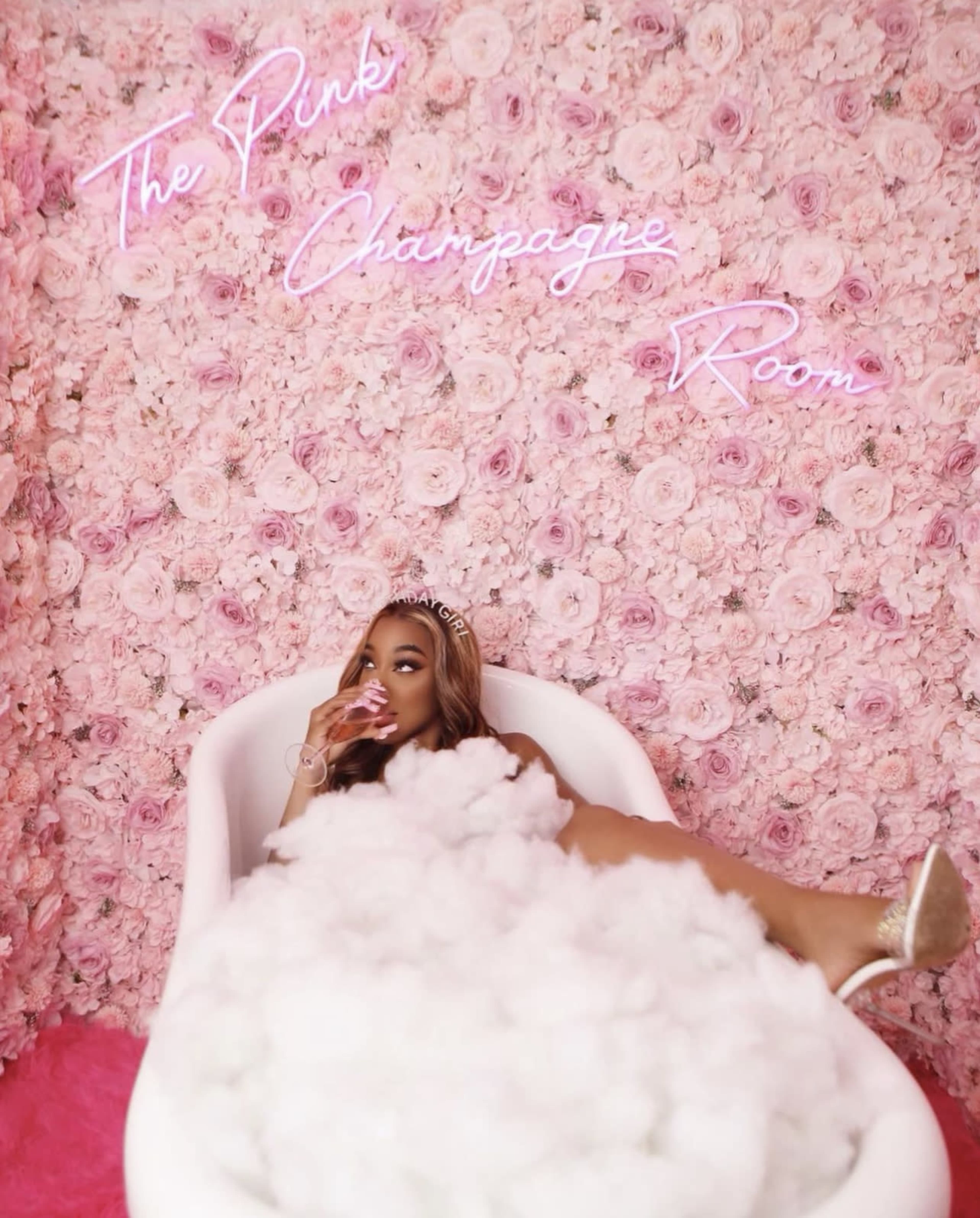 A woman lounges in a bathtub filled with clouds, set against a wall of pink roses with the words "The Pink Champagne Room" illuminated in neon.