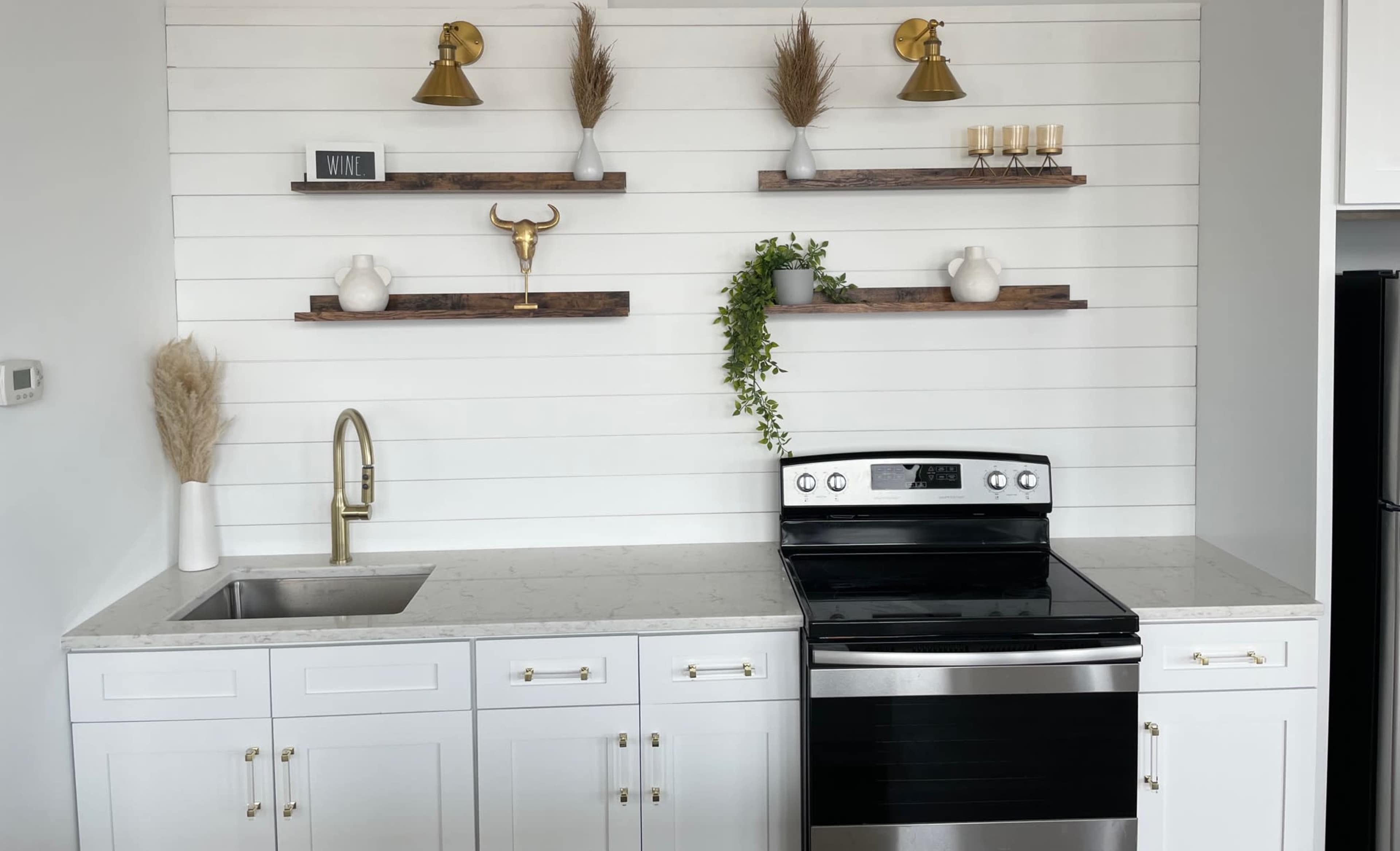 A modern kitchen features a black stove, white cabinetry, a sink with a gold faucet, and decorative shelves above with plants and accessories.