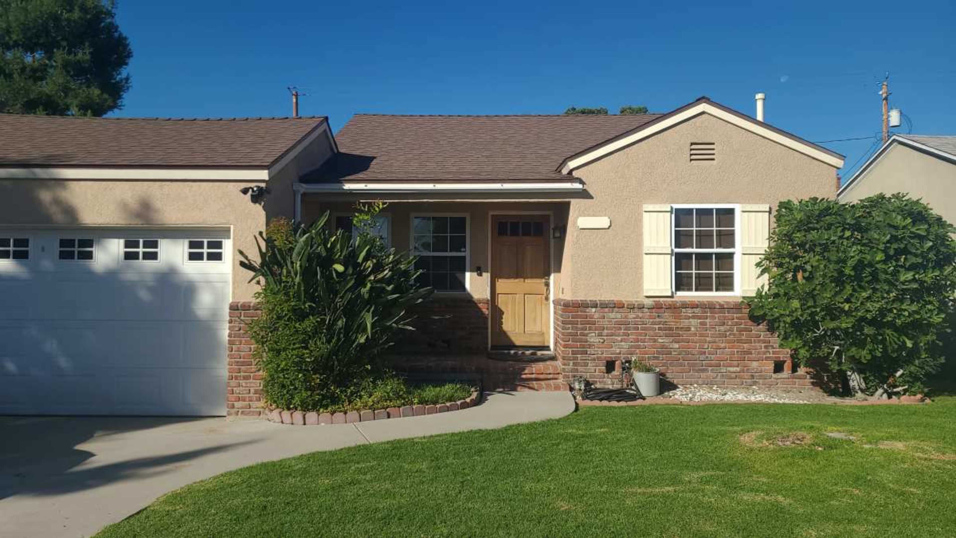 The image shows a single-story house with a brick and stucco exterior, a front yard with grass, and a driveway leading to a garage.