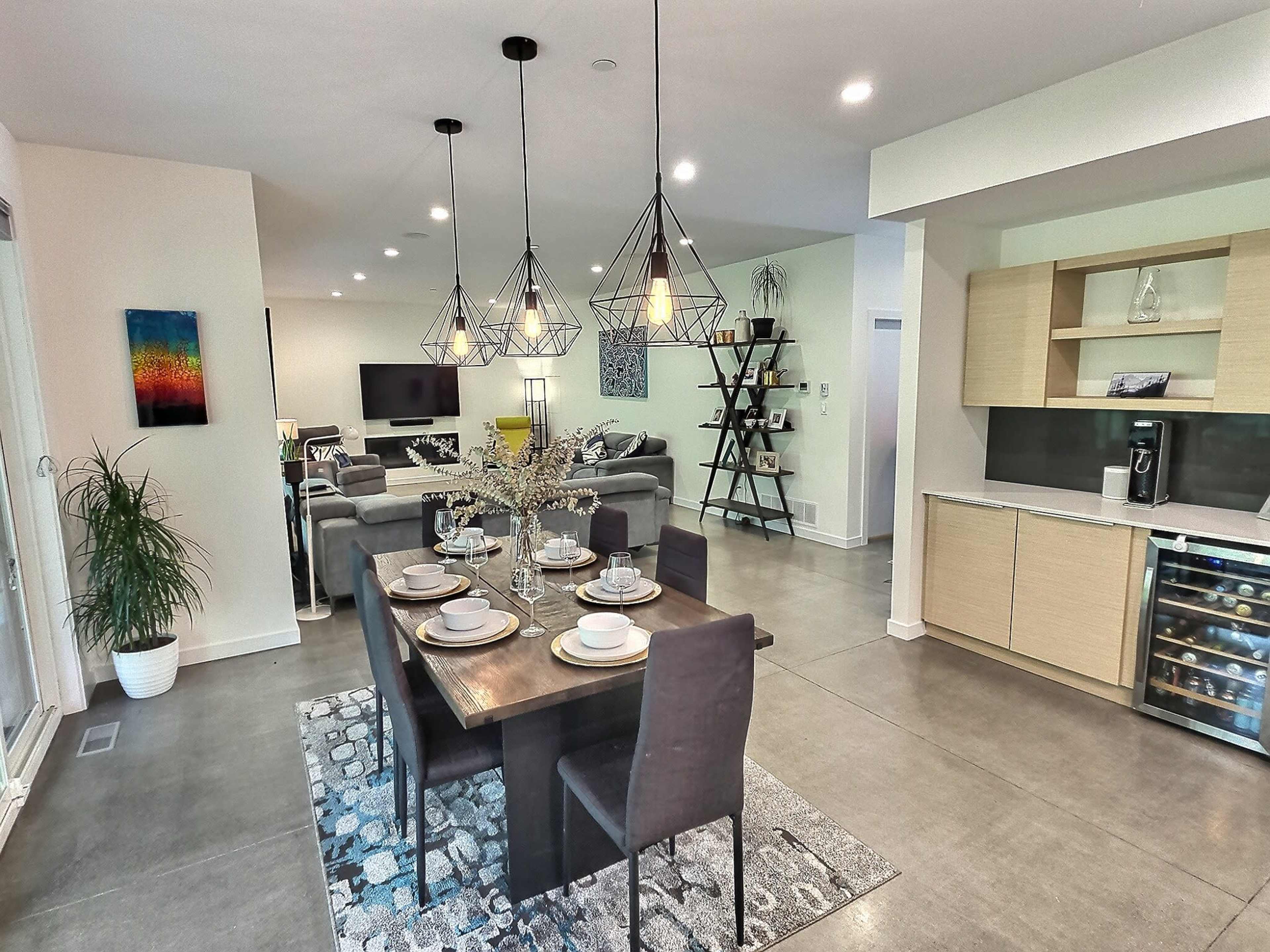 A modern open-concept living and dining area featuring a dining table set for six, contemporary lighting fixtures, and a comfortable seating area with a television.