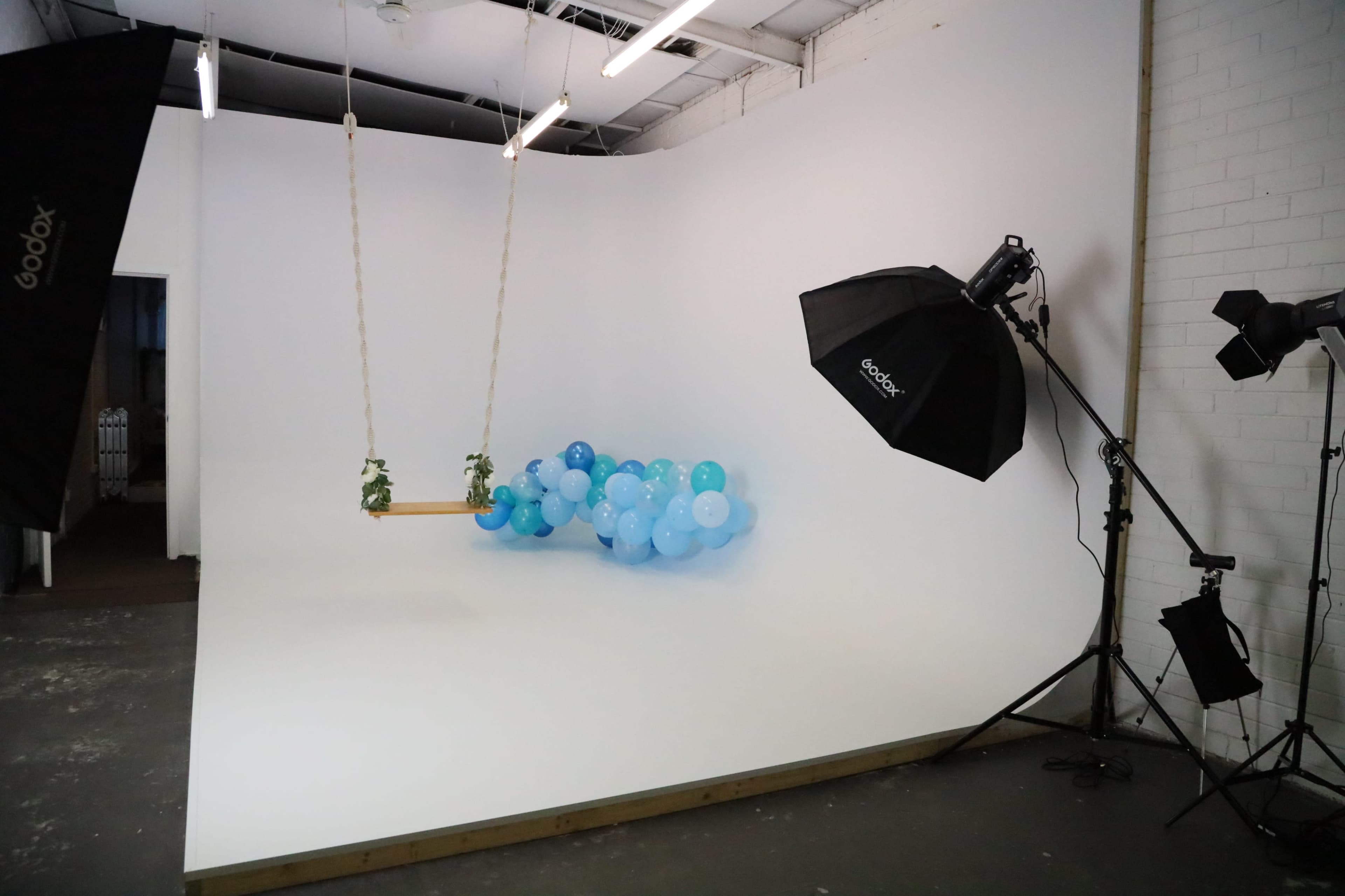 A minimalist studio setup with a white backdrop, a wooden swing with plants, and clusters of blue balloons on one side, accompanied by lighting equipment.