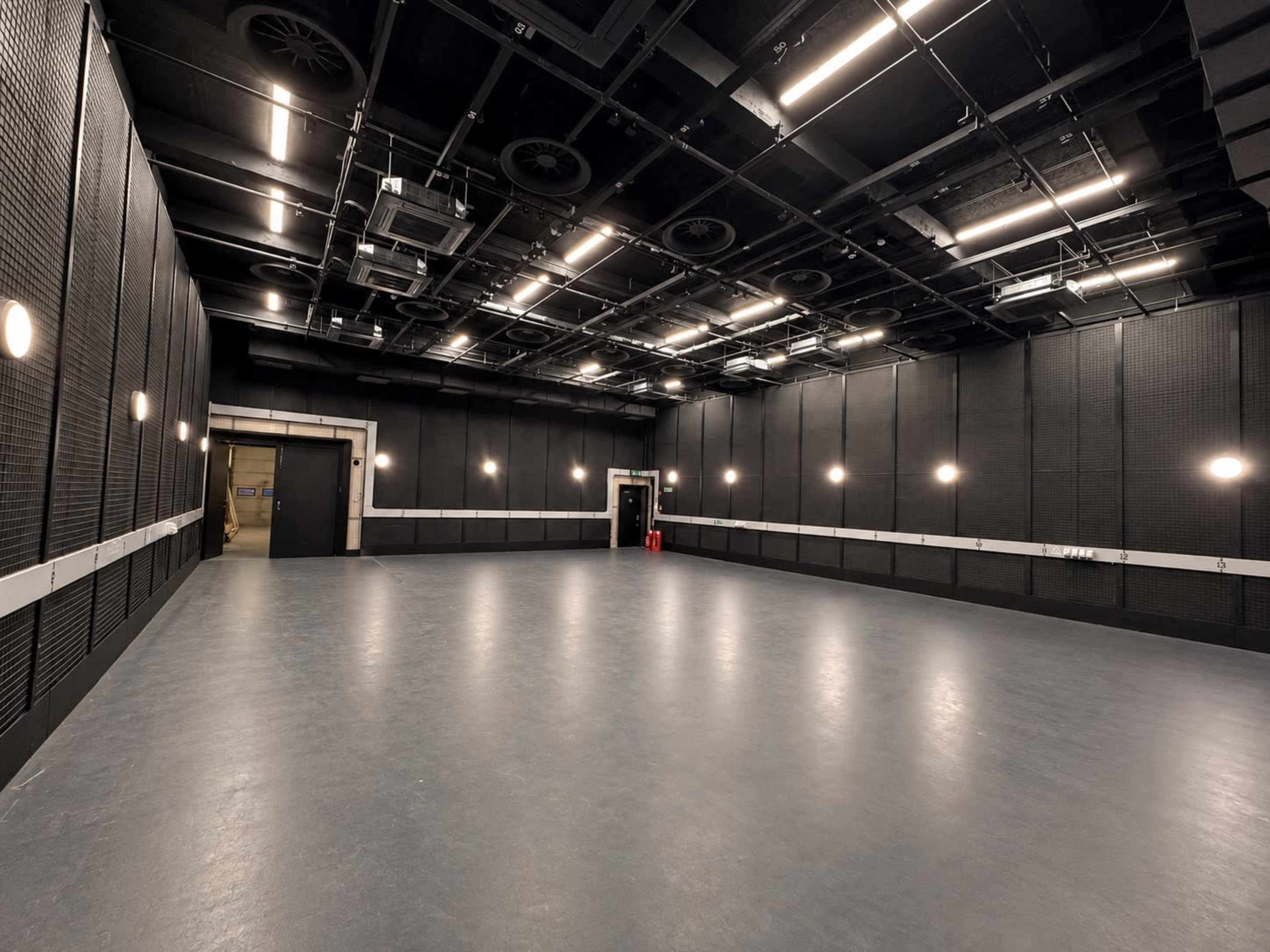 The image shows a large, empty studio space with dark walls and ceiling fixtures, featuring a smooth gray floor and uniform lighting along the walls.