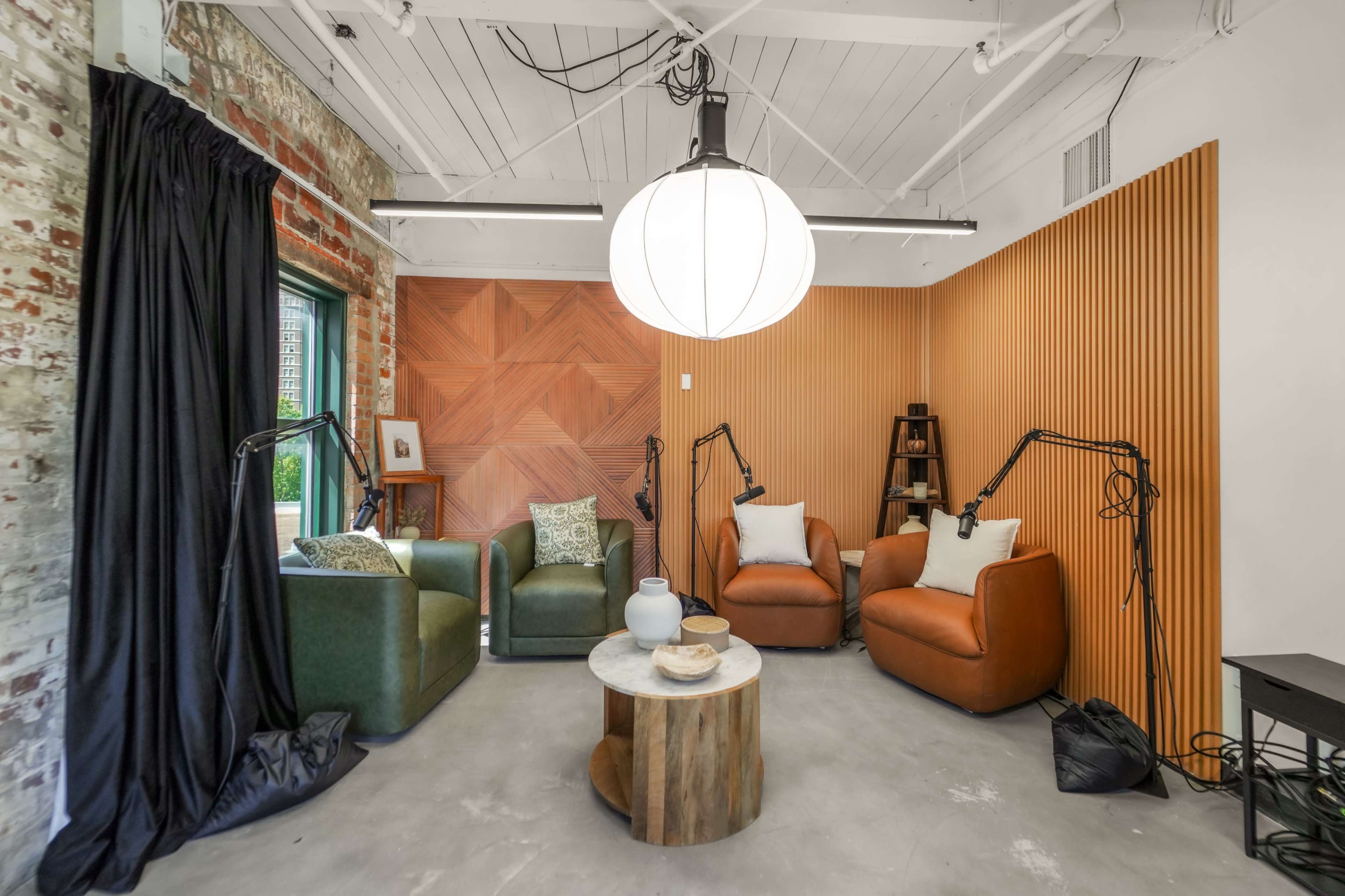 The image shows a modern conversation area with two armchairs, a sofa, a round wooden coffee table, and decorative elements against a backdrop of wooden panels and exposed brick.