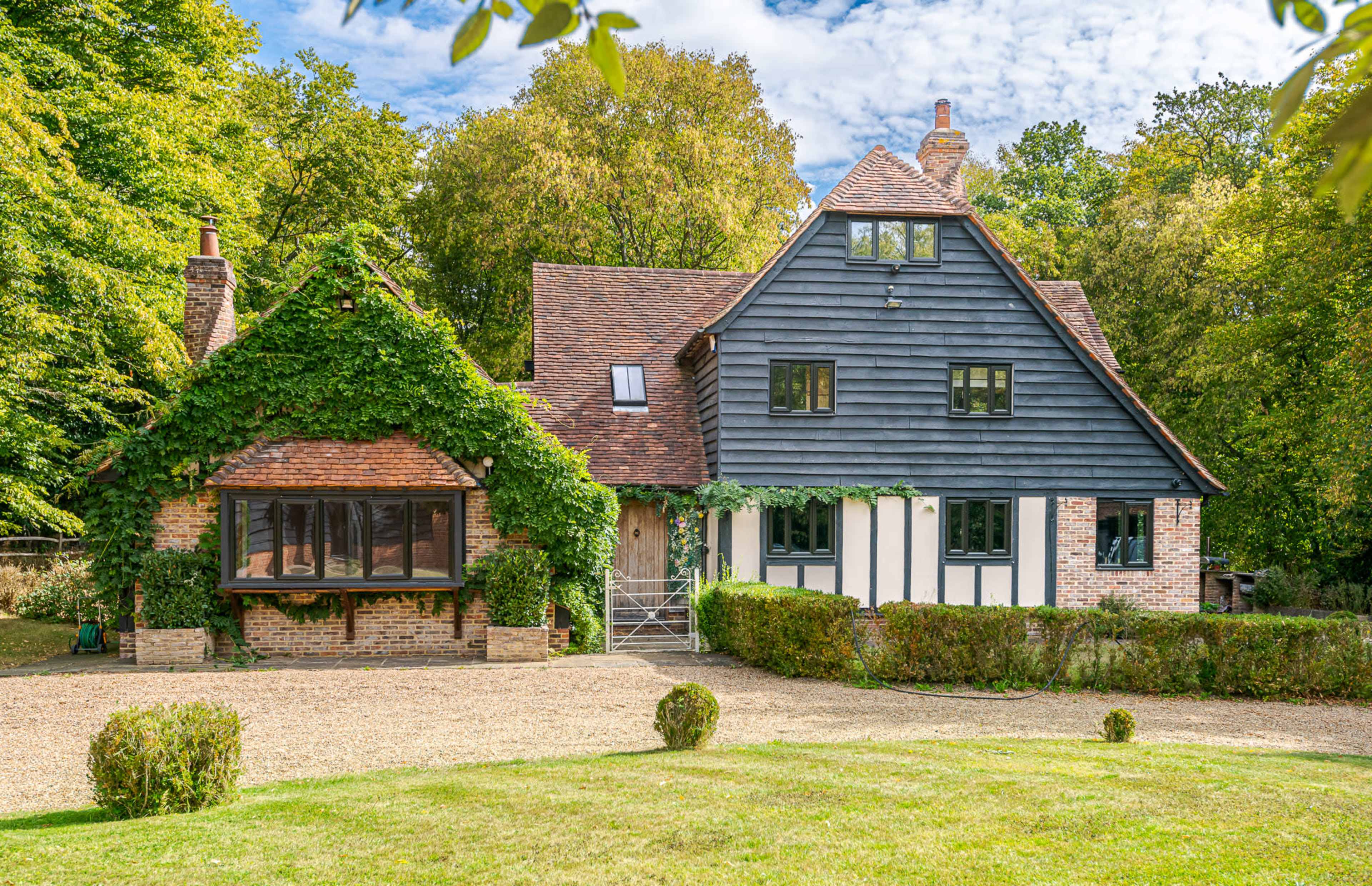 A two-story house with dark wooden siding and a green, vine-covered entrance is situated within a lush garden surrounded by trees.