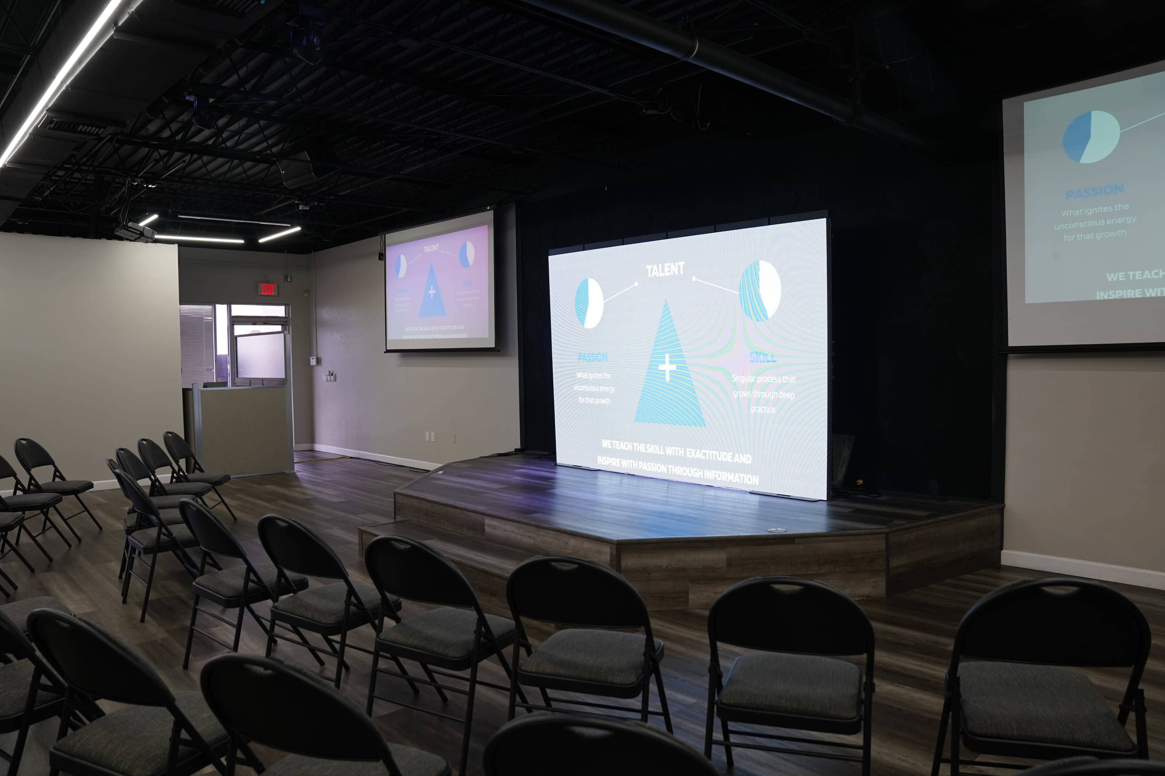 The image shows a modern presentation space with a multi-screen display at the front, surrounded by rows of black chairs for an audience.