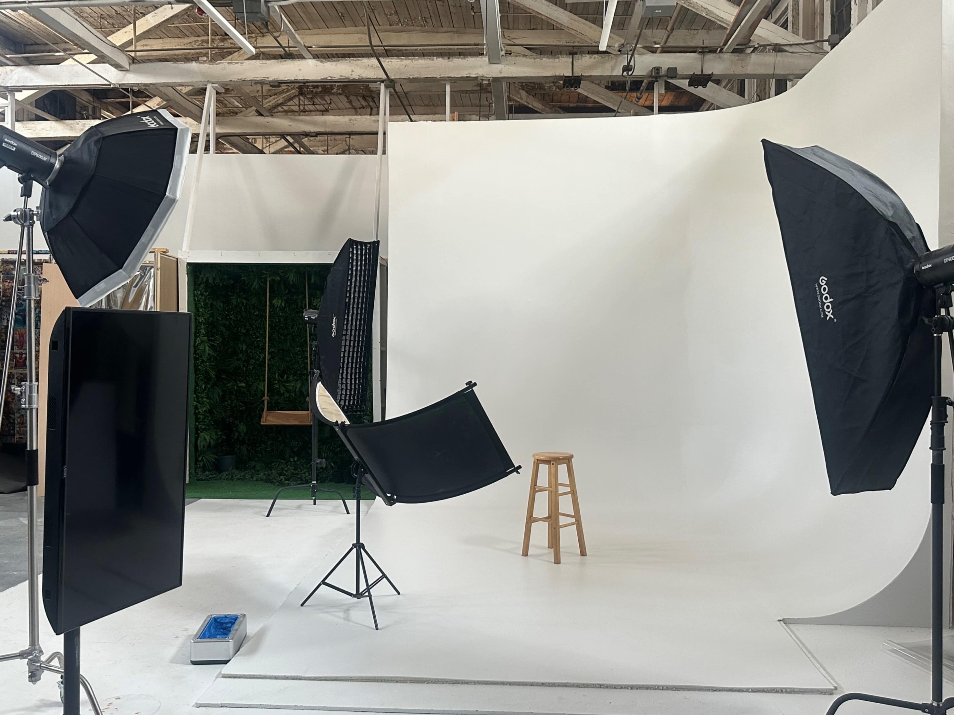 A photography studio features a white backdrop, a wooden stool, and various lighting equipment arranged around the space.