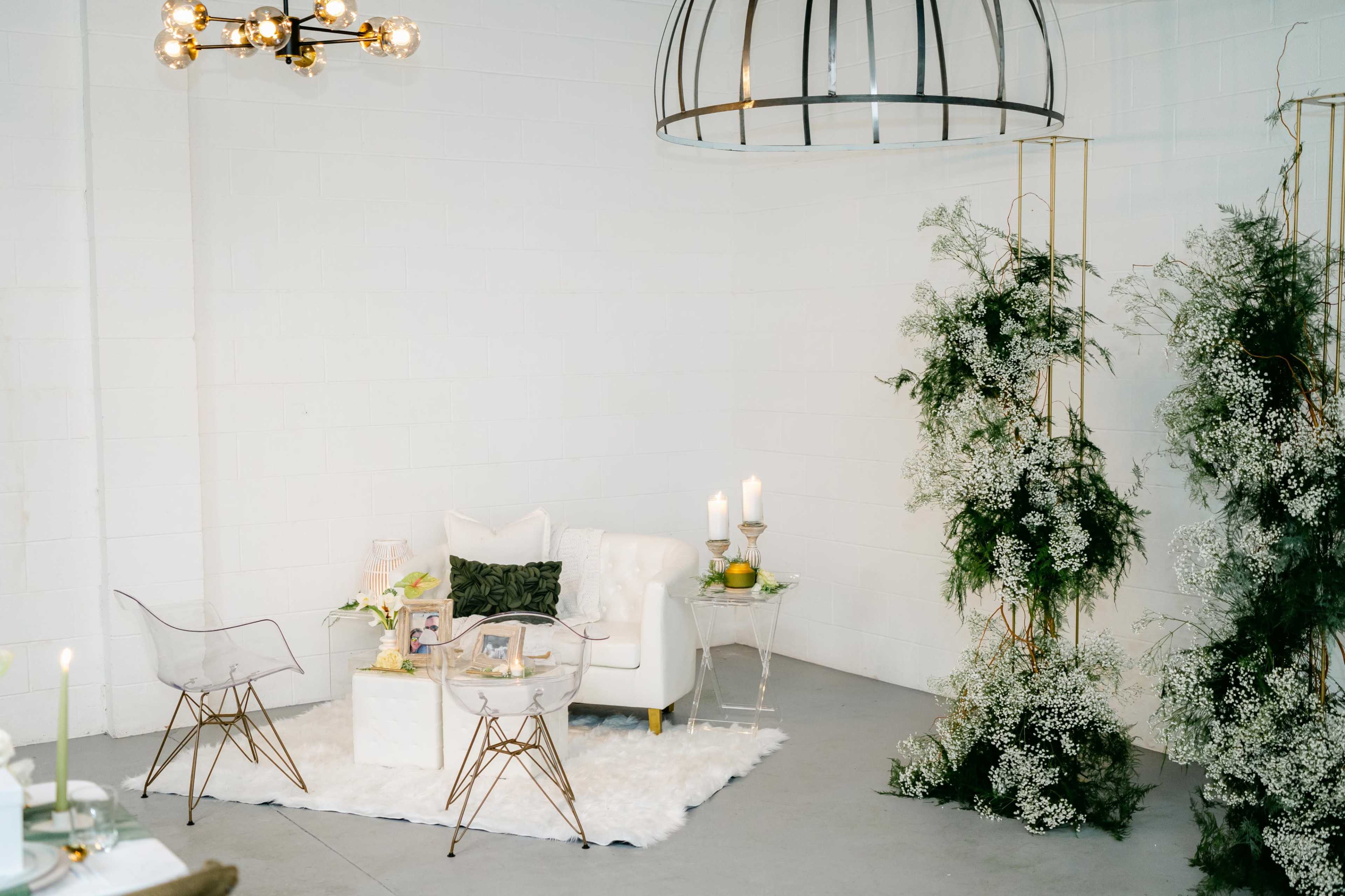 A minimalist lounge area featuring a white tufted sofa, a clear coffee table, and decorative arrangements of greenery, set against a plain white wall.