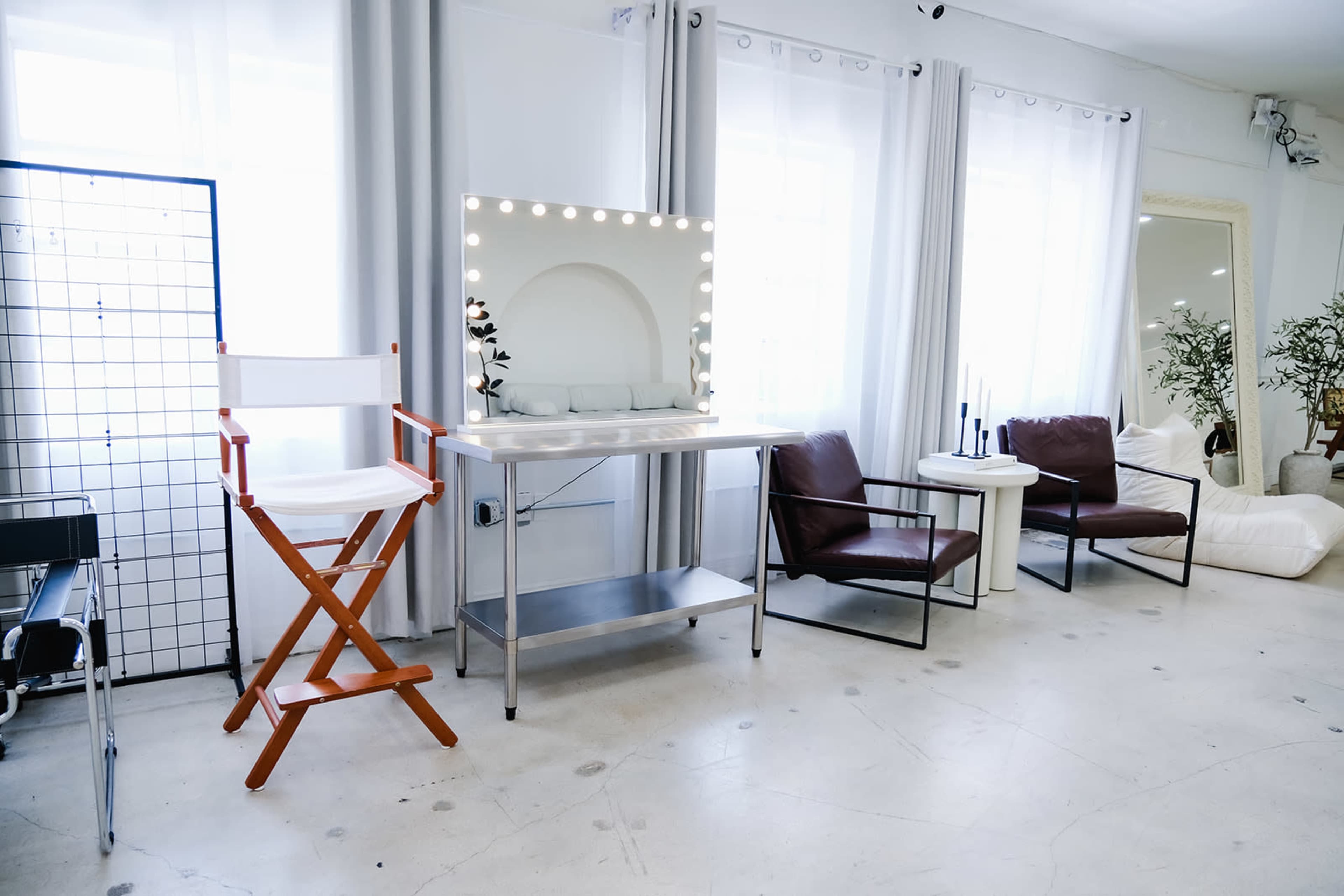 The image depicts a bright, modern room featuring a makeup station with a lighted mirror, a director's chair, and several contemporary seating options.