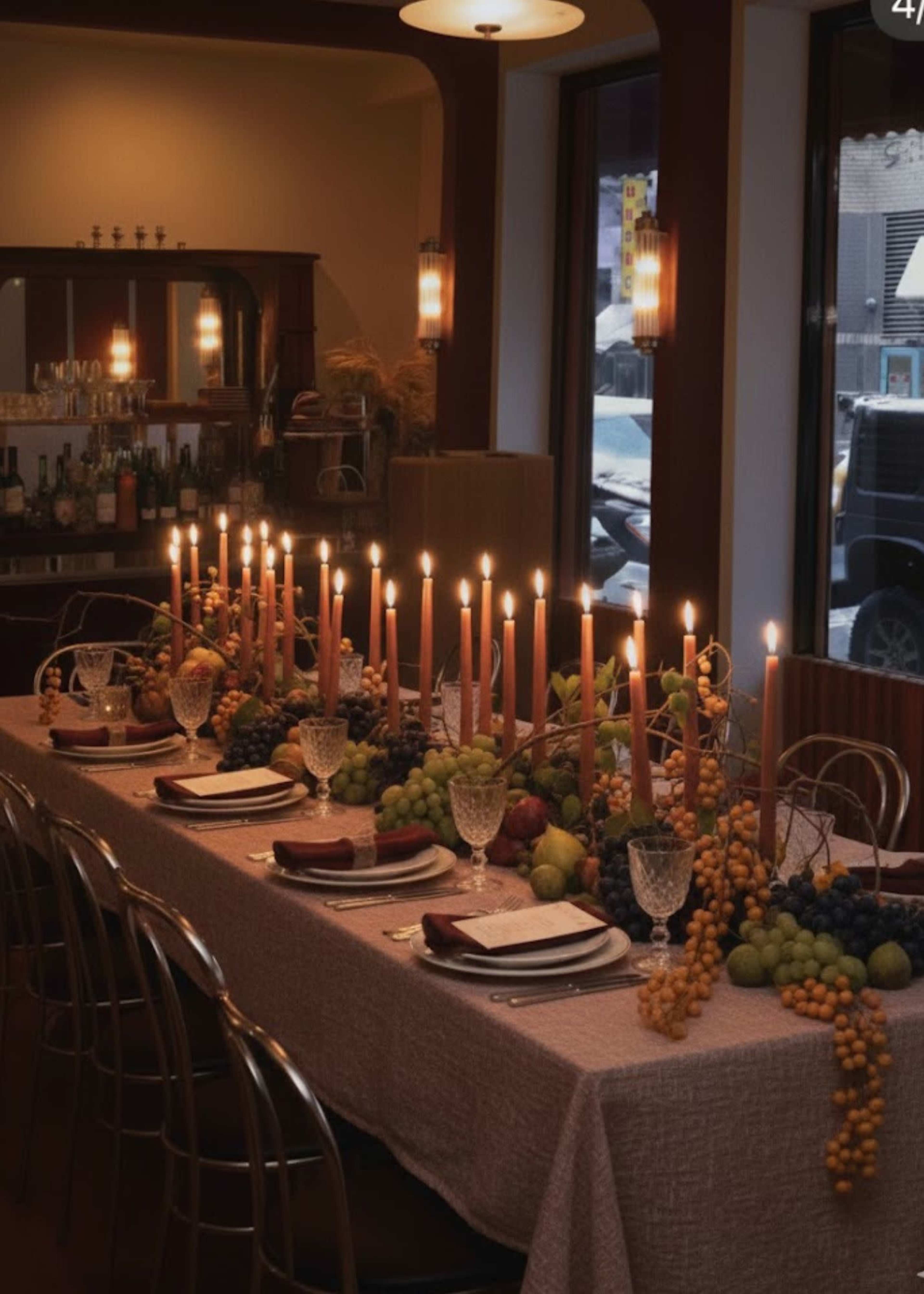 A long dining table is set with plates, glasses, and candles, adorned with various fruits arranged along the table's center.