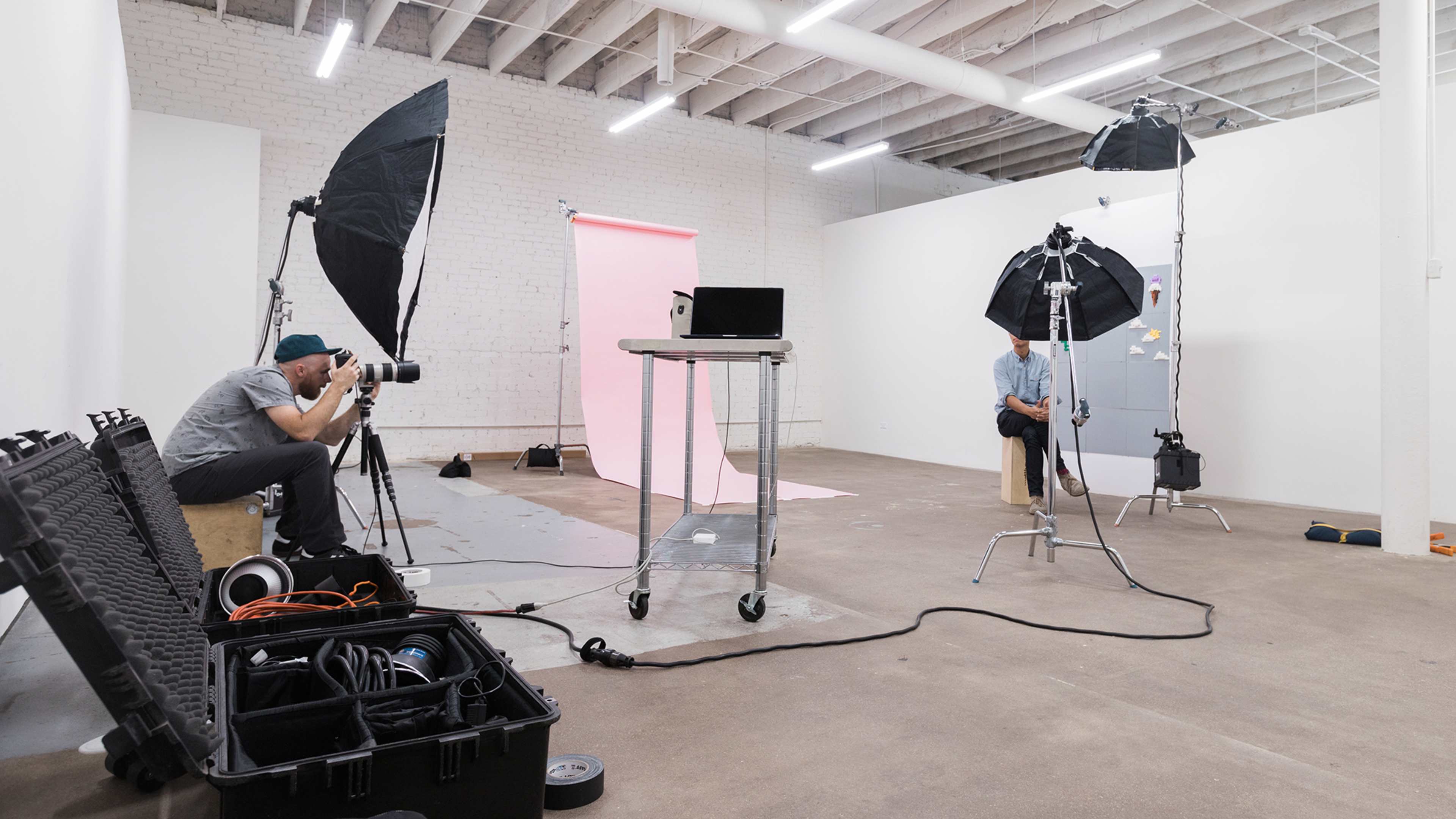 A photographer is setting up a shoot in a studio with multiple lights and a laptop, while a person sits on a stool against a pink backdrop.