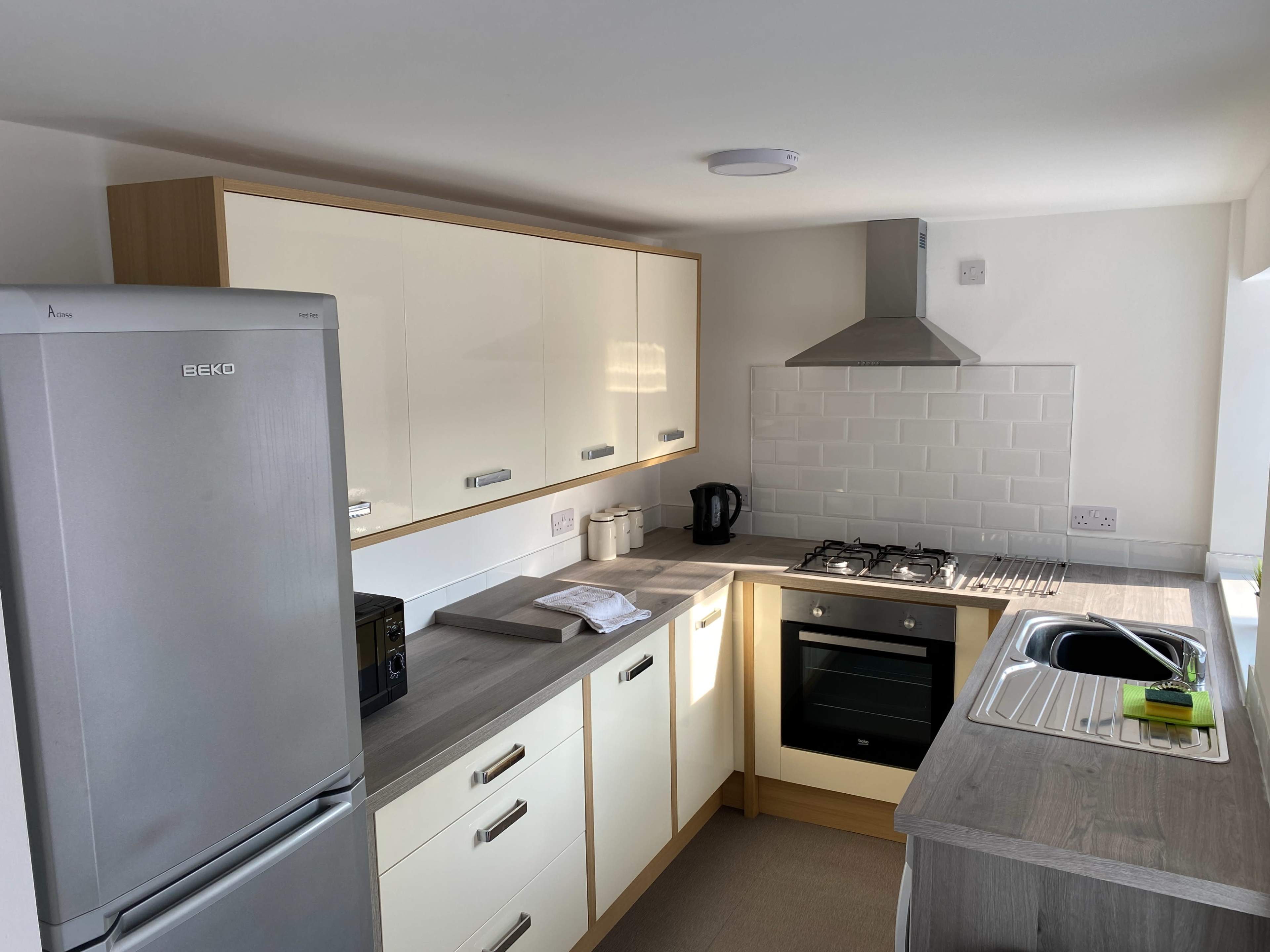 The image shows a modern kitchen with a silver refrigerator, a gas stove, an oven, and a sink, featuring light-colored cabinetry and a tiled backsplash.