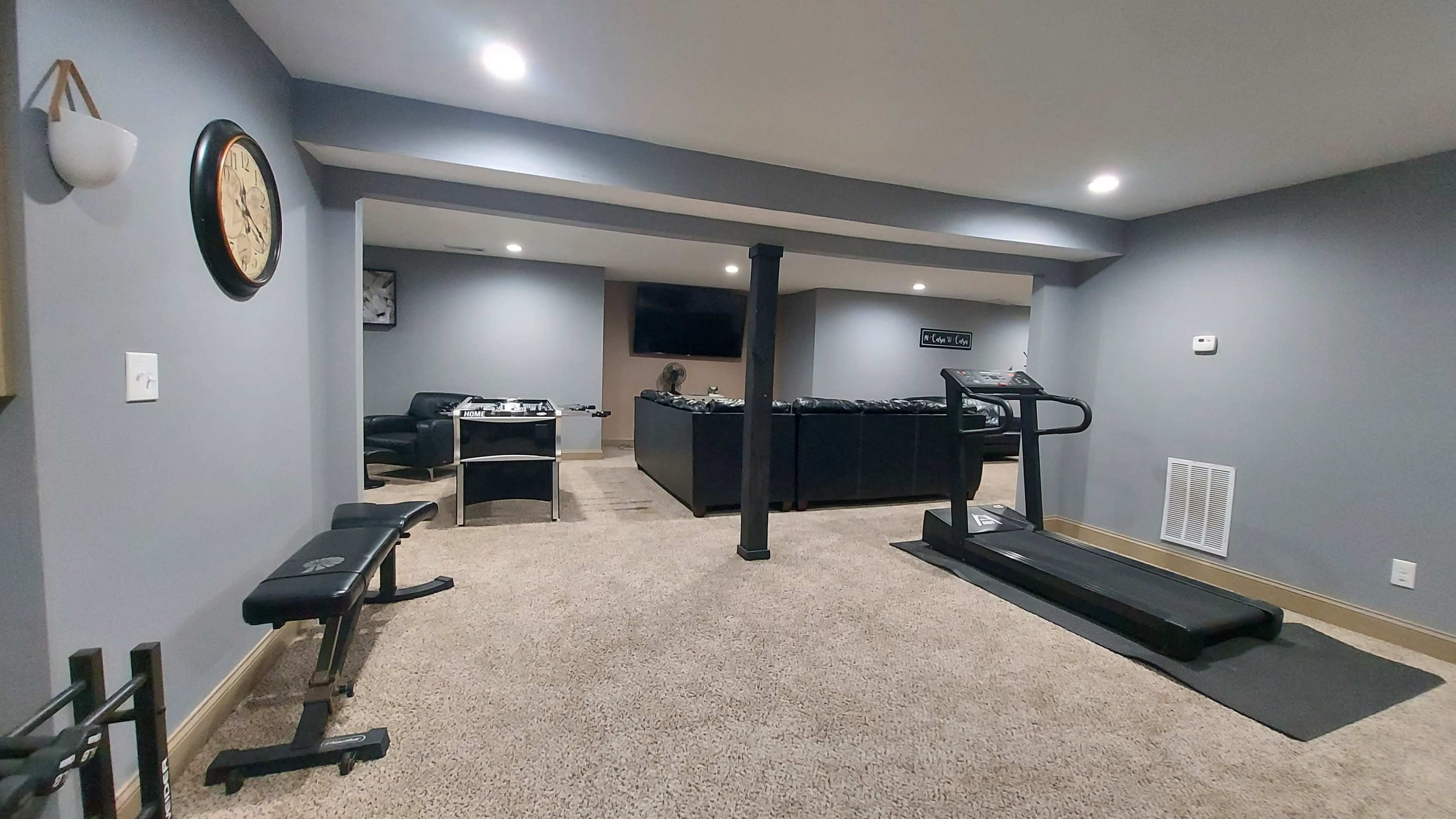 A home gym with carpeted flooring, a treadmill, a weight bench, and a desk area in a well-lit basement.