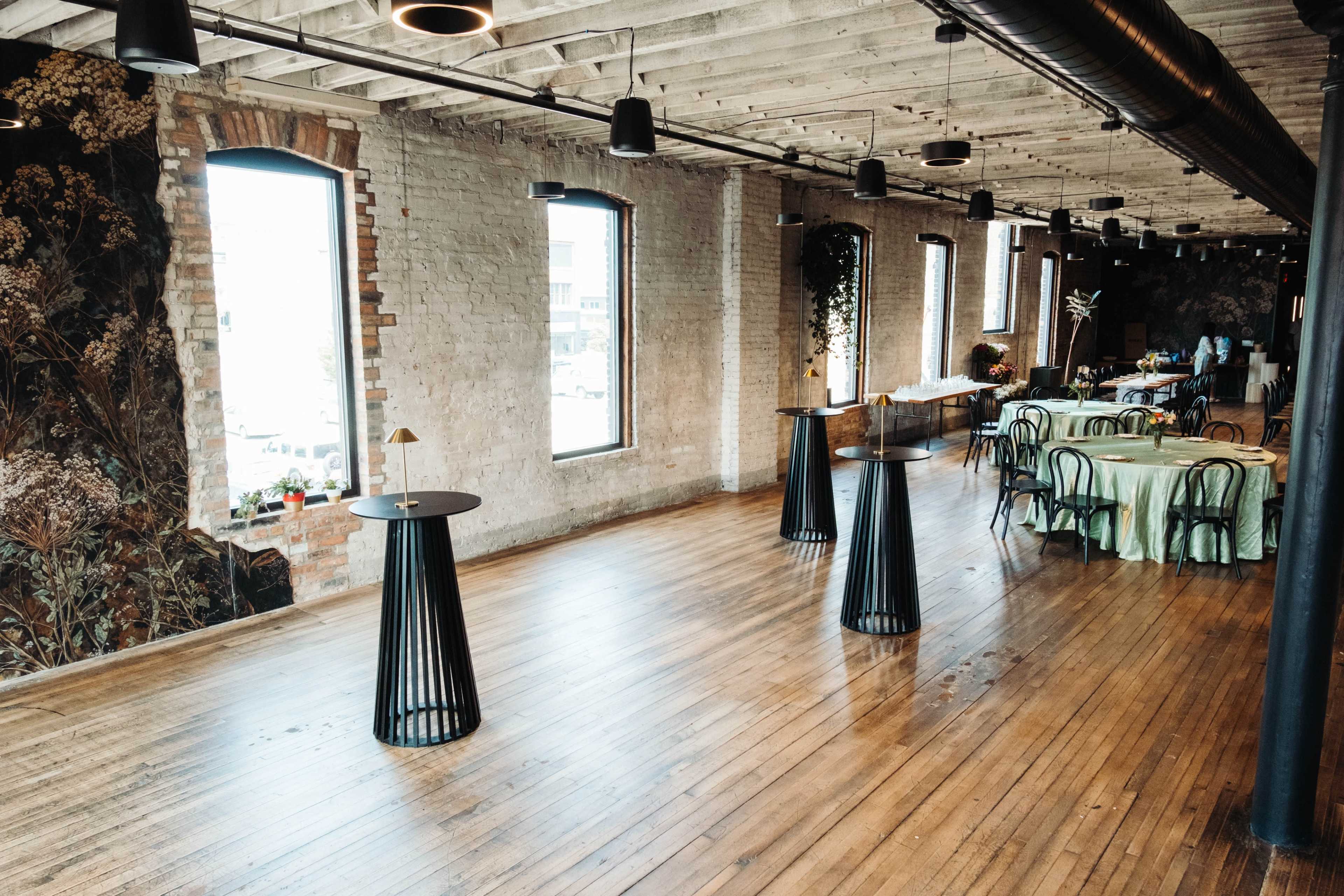 The image shows a spacious event venue with wooden floors, industrial-style lighting, and large windows, featuring several black tables and decorated dining areas.