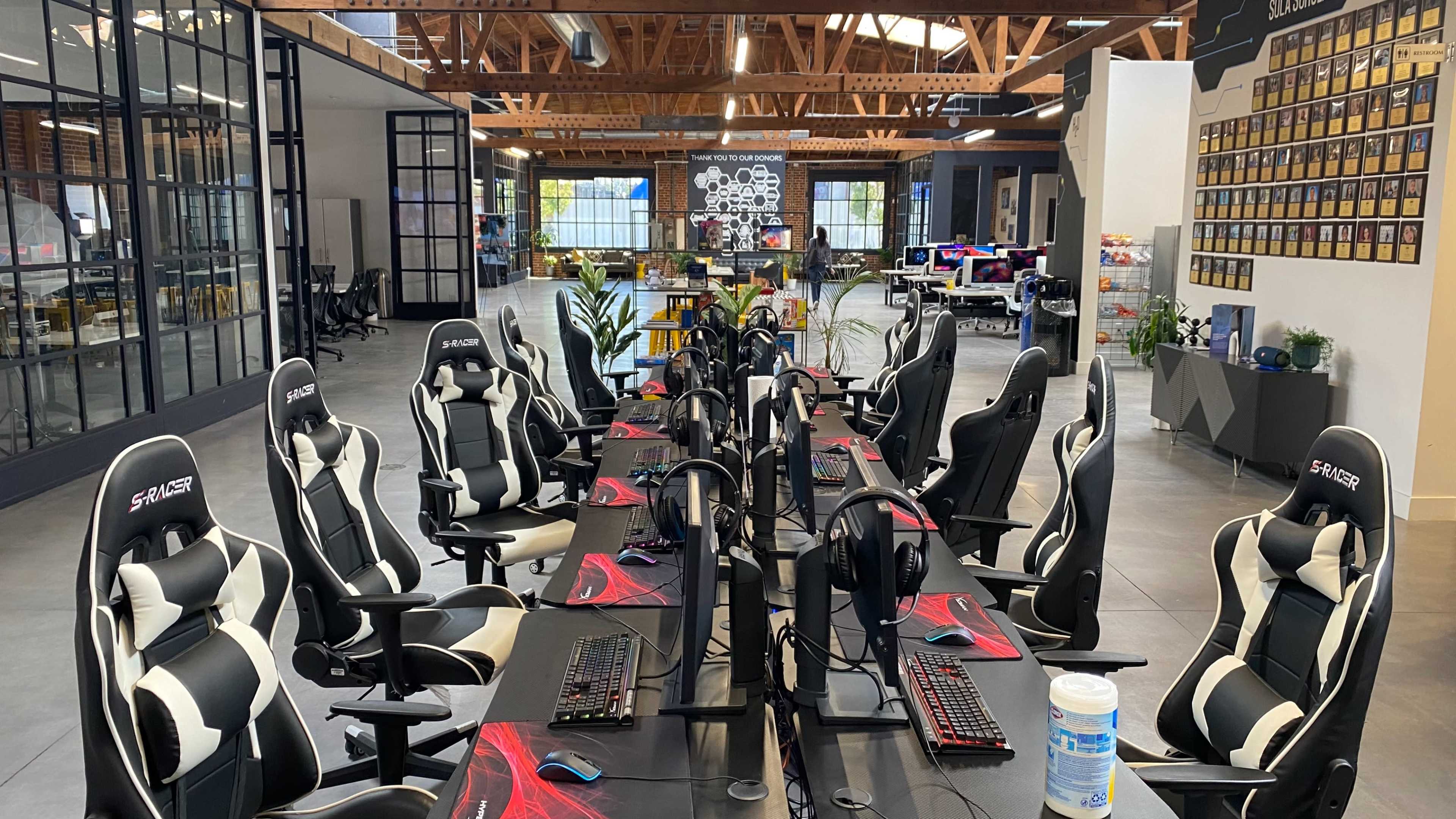 A modern open office space with several gaming desks equipped with monitors and gaming chairs arranged in a row.