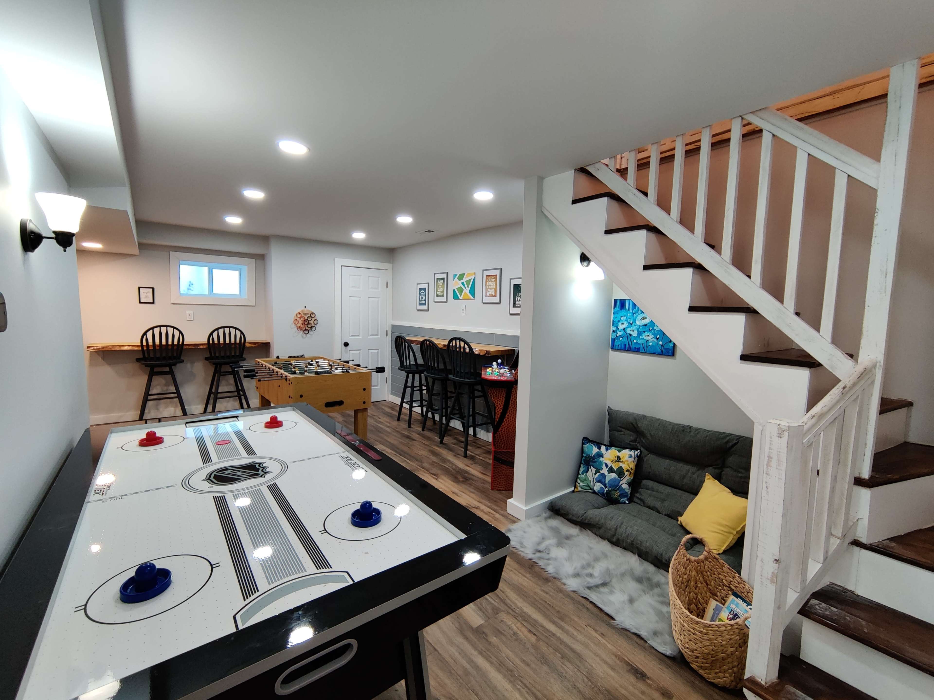 A game room features an air hockey table, seating areas with chairs and a couch, and stairs leading to another level.
