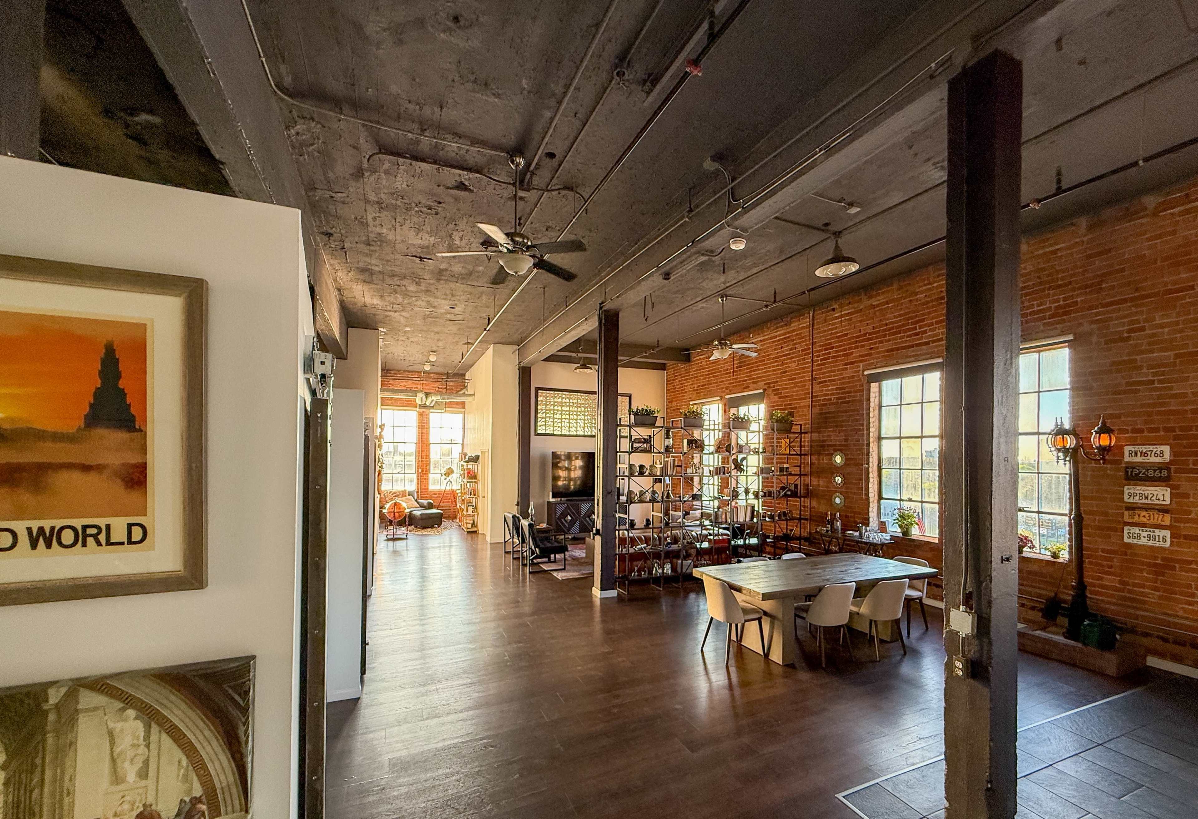 The image shows an industrial-style interior space with exposed brick walls, wooden flooring, and a dining area beside a large window.