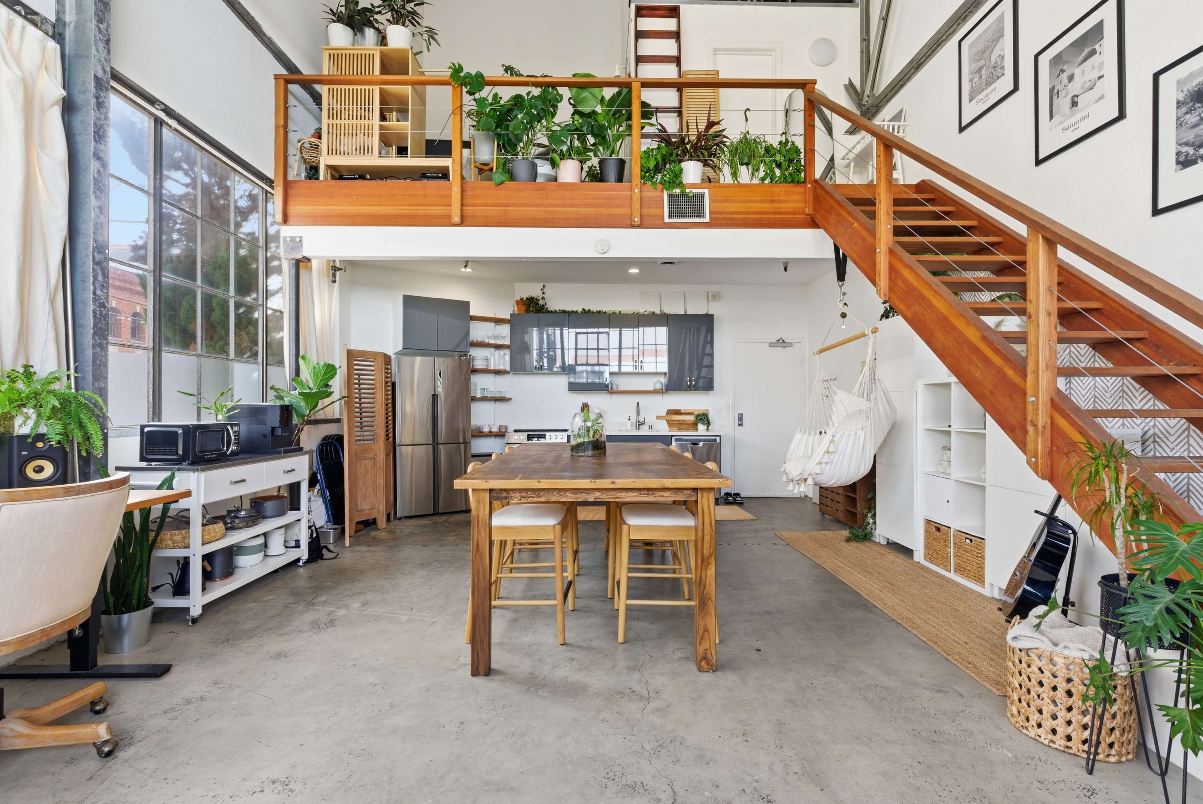 The image shows a spacious, modern loft with a kitchen area, wooden dining table, and a mezzanine level surrounded by various plants.